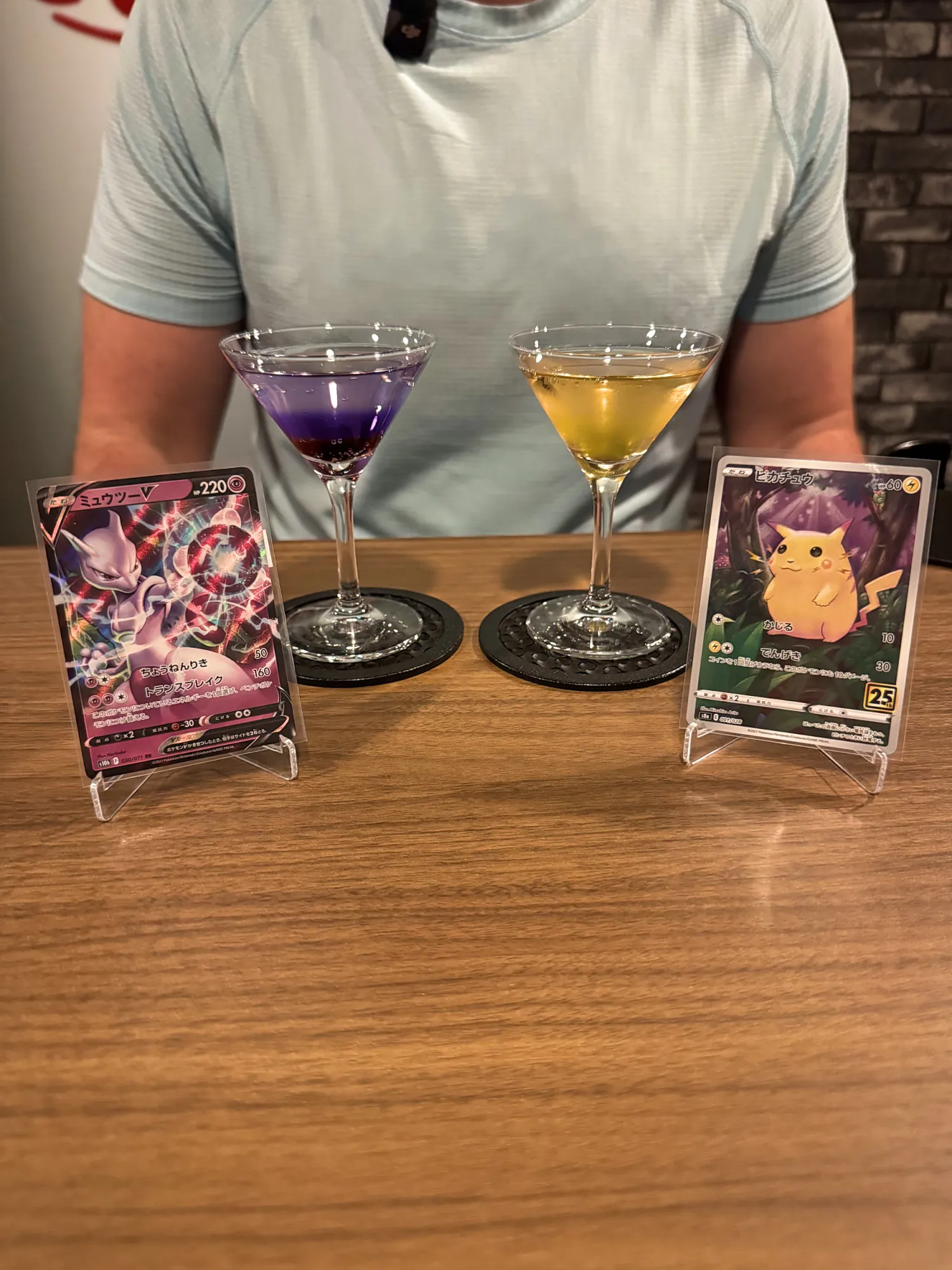 Two drinks at a Pokémon card cocktail bar in Tokyo, a purple one beside a Mewtwo V card and a yellow one beside a Pikachu card, each card on a small stand.