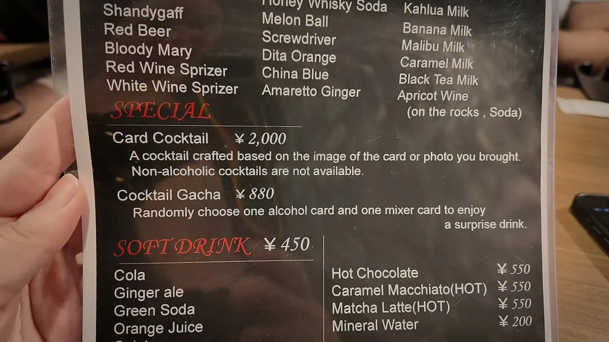 A hand holding FUN's English drinks menu, listing the ¥2,000 Card Cocktail made from the card or photo you bring, a ¥880 Cocktail Gacha, and ¥450 soft drinks.