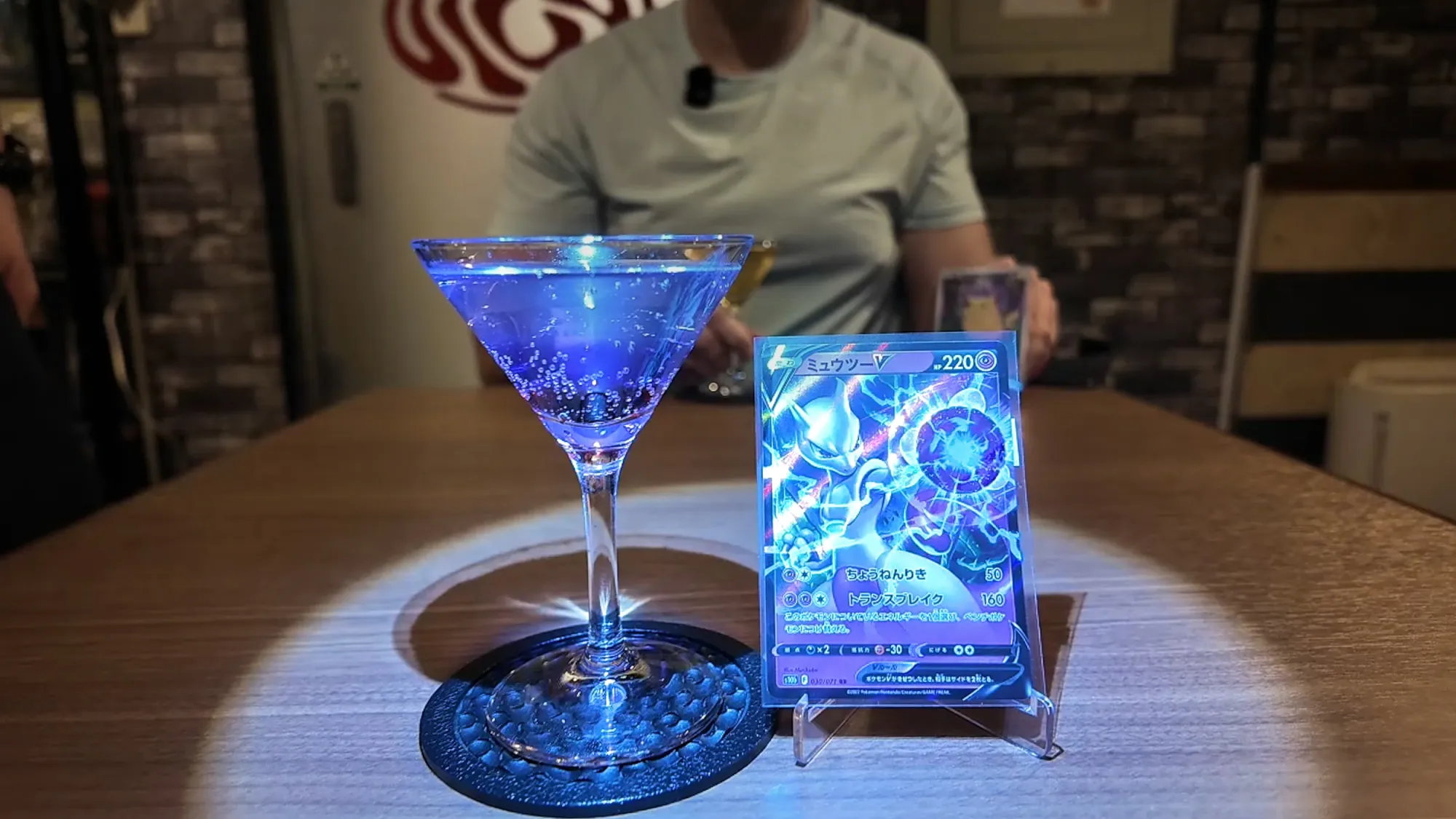 A glowing blue-violet cocktail in a martini glass beside a Mewtwo V Pokémon card on a stand, lit by a spotlight on a wooden table at FUN in Tokyo.