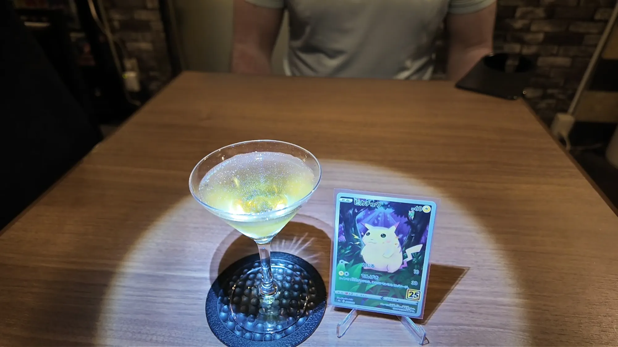 A pale cocktail in a martini glass beside a Pikachu Pokémon card on a clear stand, under a spotlight on the table at FUN in Tokyo.