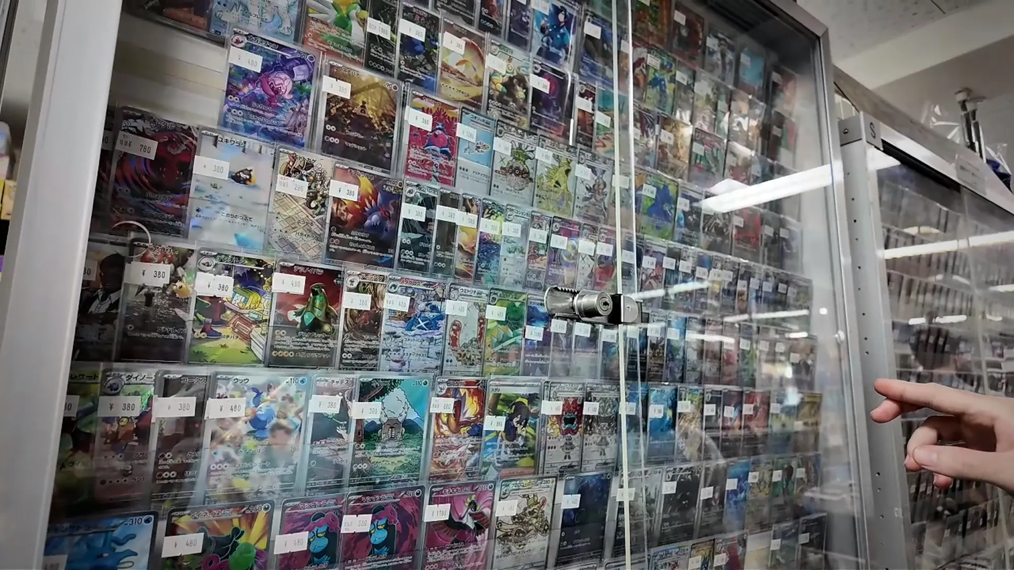 A finger pointing at rows of single Pokémon cards in a glass display case at a Tokyo card shop.