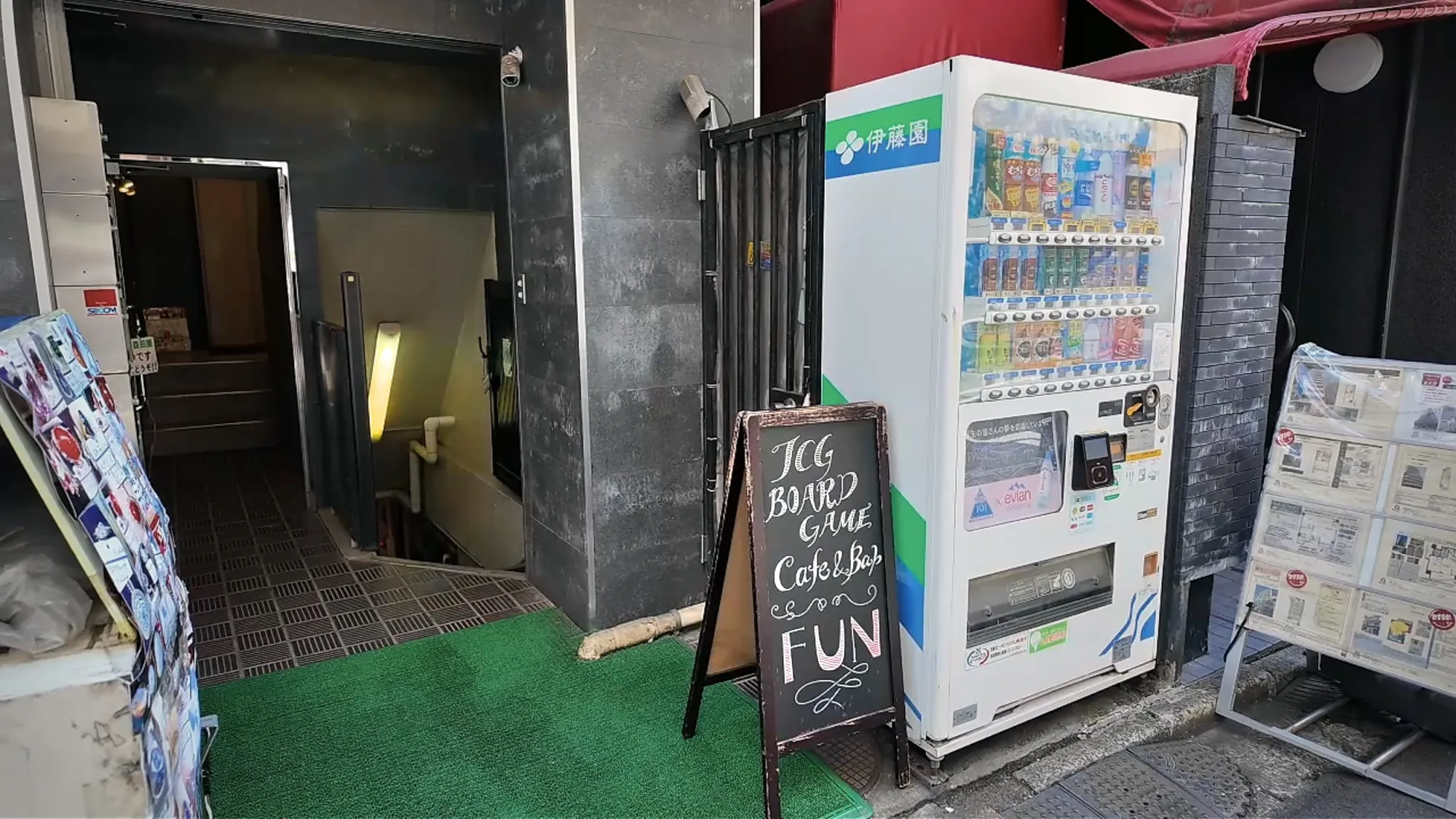 The chalkboard sign for TCG Board Game Cafe & Bar FUN beside a drinks vending machine, next to the stairs down to the basement bar in Ueno.