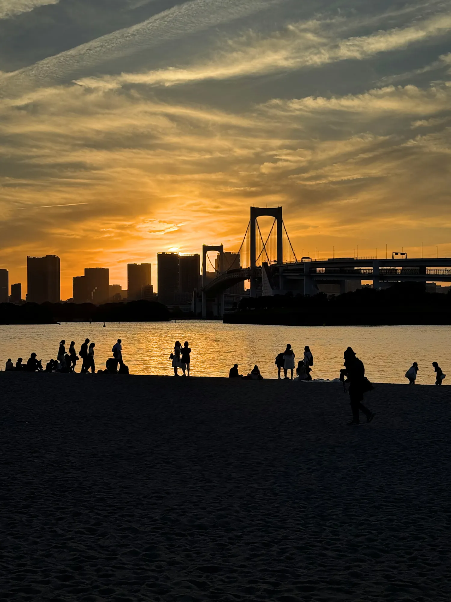 The Rainbow Bridge and the Tokyo skyline silhouetted against an orange sunset over Tokyo Bay, seen from the beach at Odaiba, Japan.