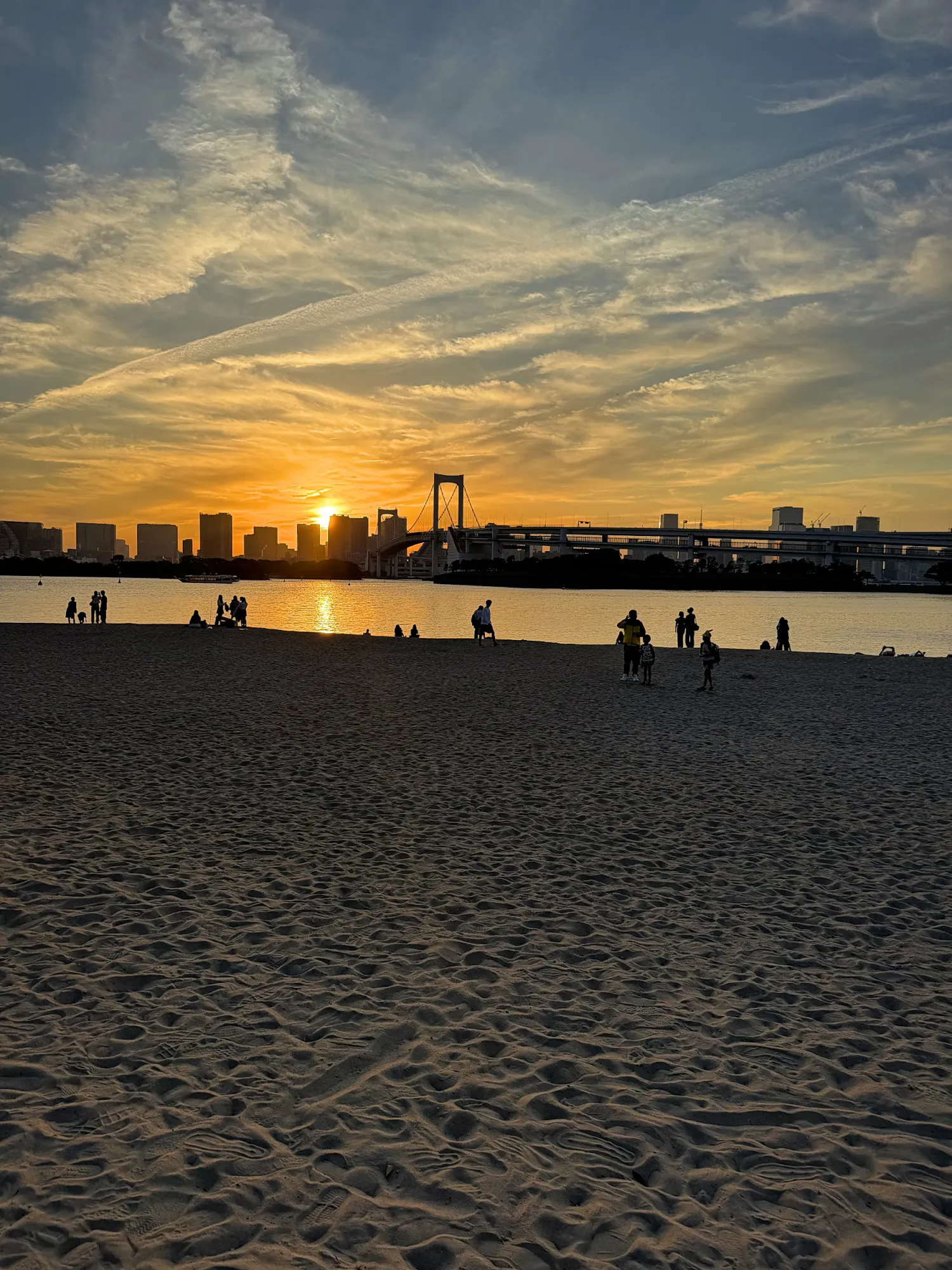 The sun setting behind Rainbow Bridge over Tokyo Bay, seen from the sand at Odaiba Seaside Park.