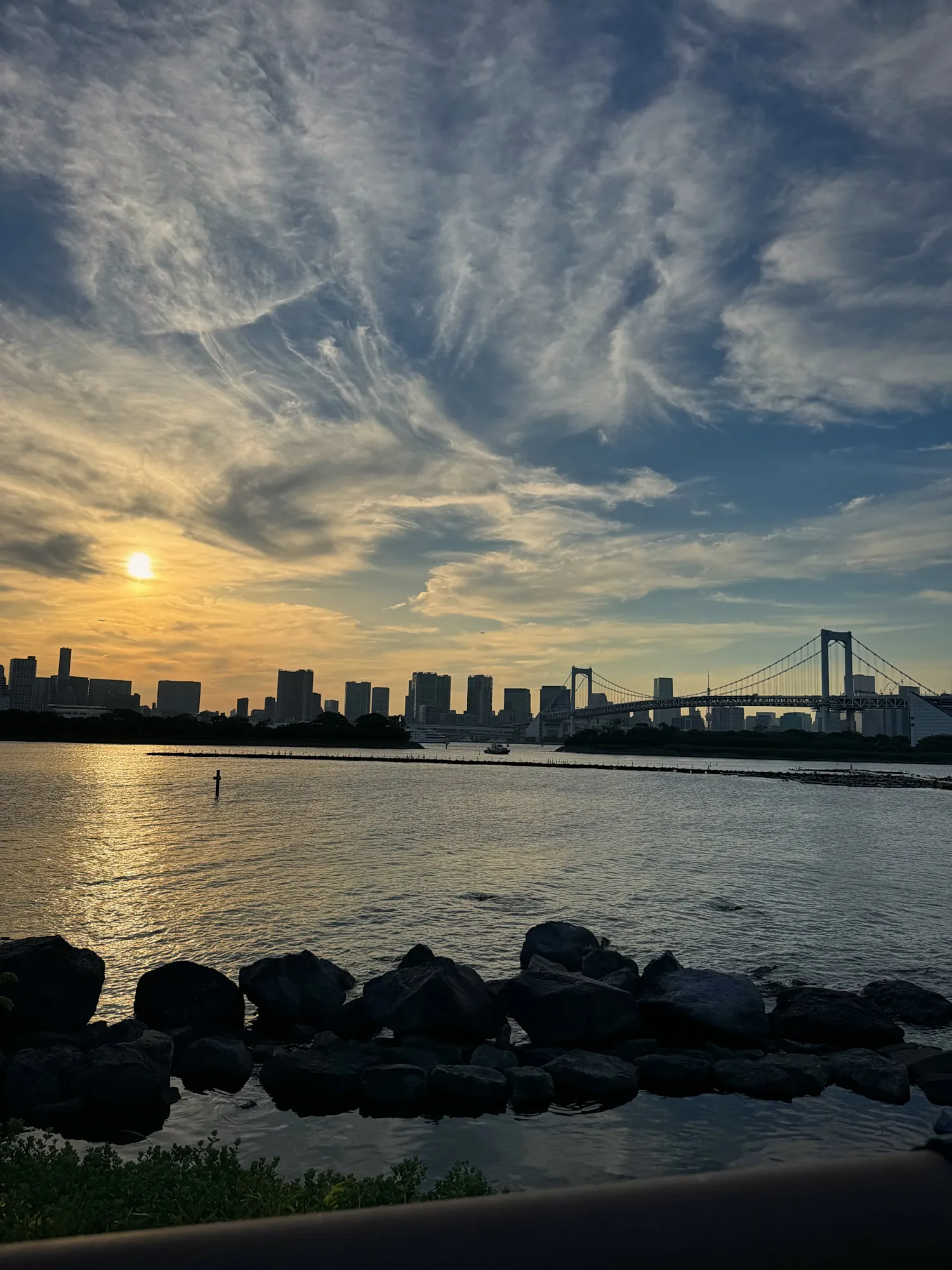 The sun setting over Tokyo Bay behind Rainbow Bridge and the city skyline, seen from the rocky shore of Odaiba, Tokyo.