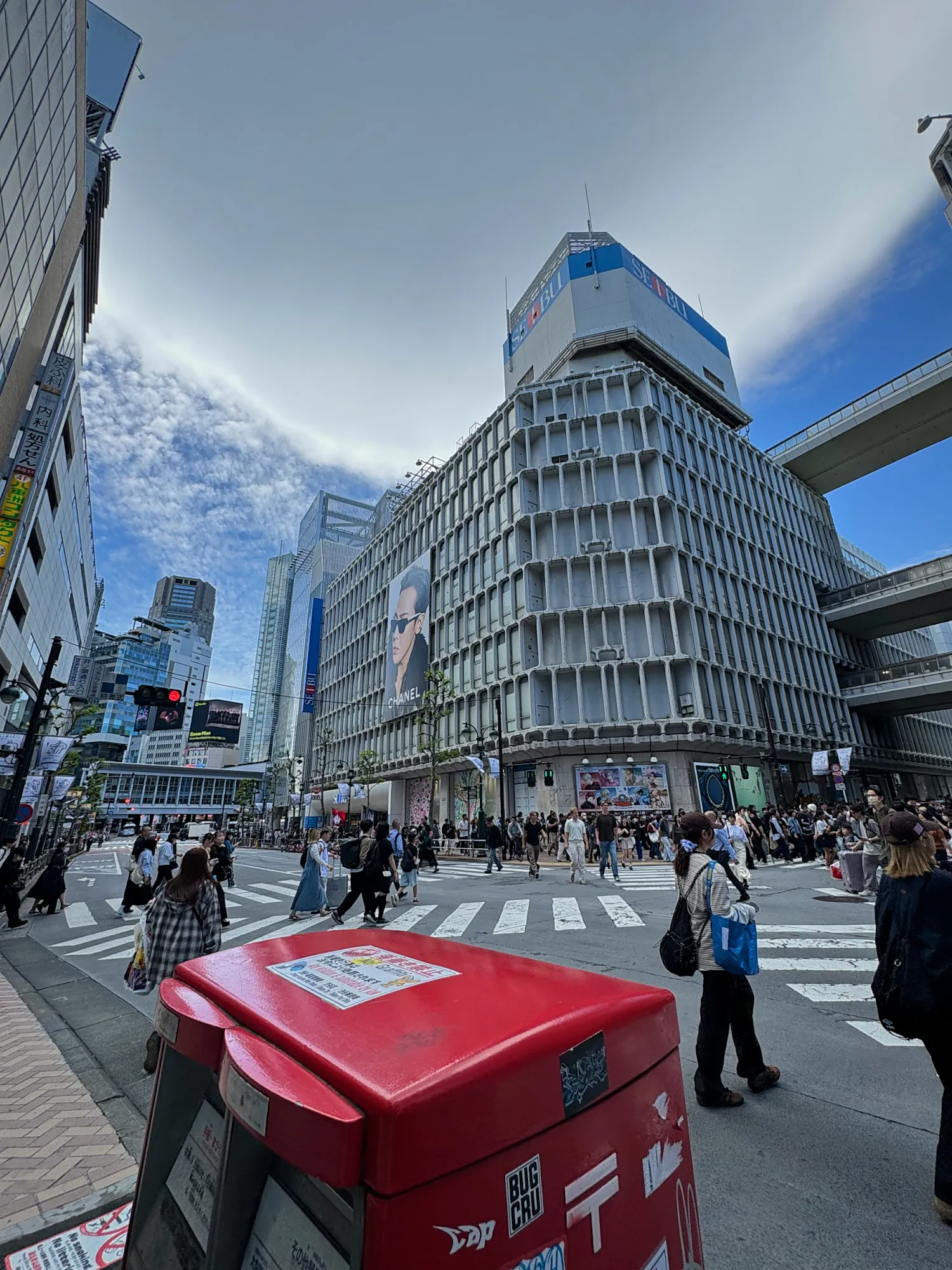 People crossing a wide street between the Seibu department store buildings in Shibuya, Tokyo, with an elevated road behind.