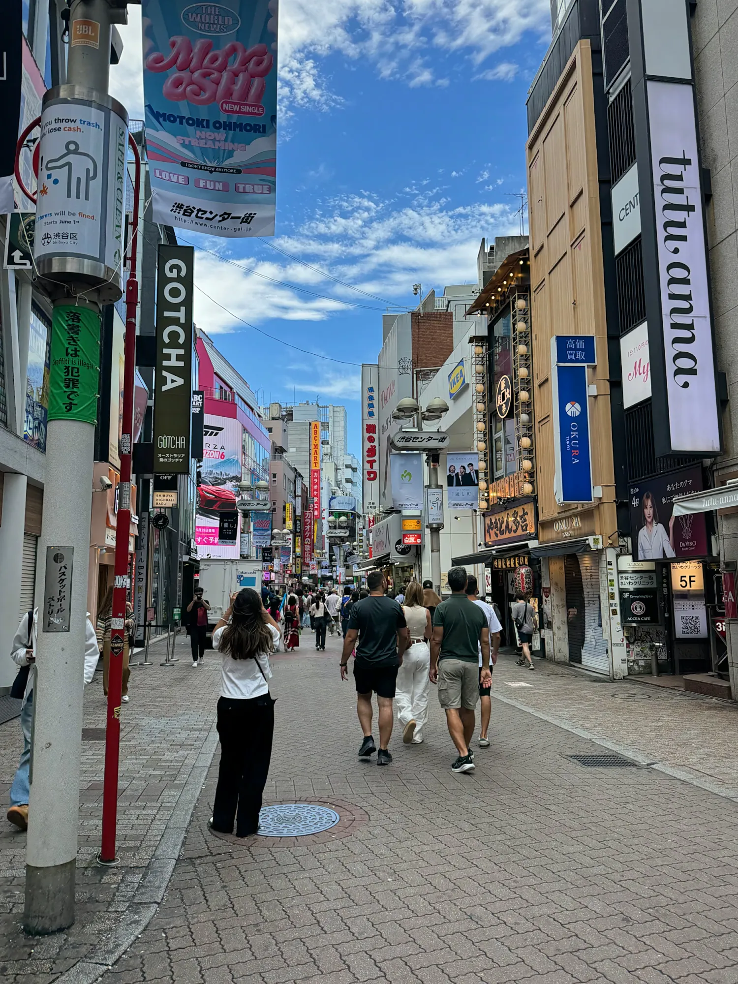 Pedestrians in everyday summer clothes walk down a busy Shibuya shopping street lined with shops, signs, and banners under a bright blue sky.