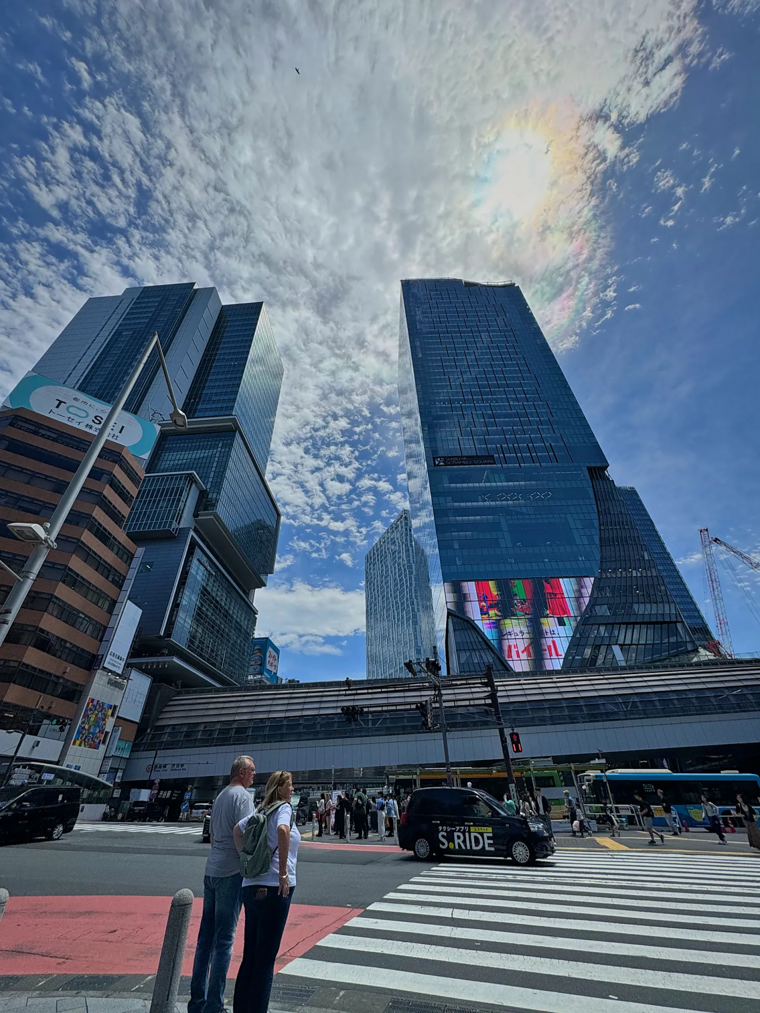 Glass skyscrapers rising above a wide crosswalk in Shibuya, Tokyo, under a blue sky with scattered clouds.