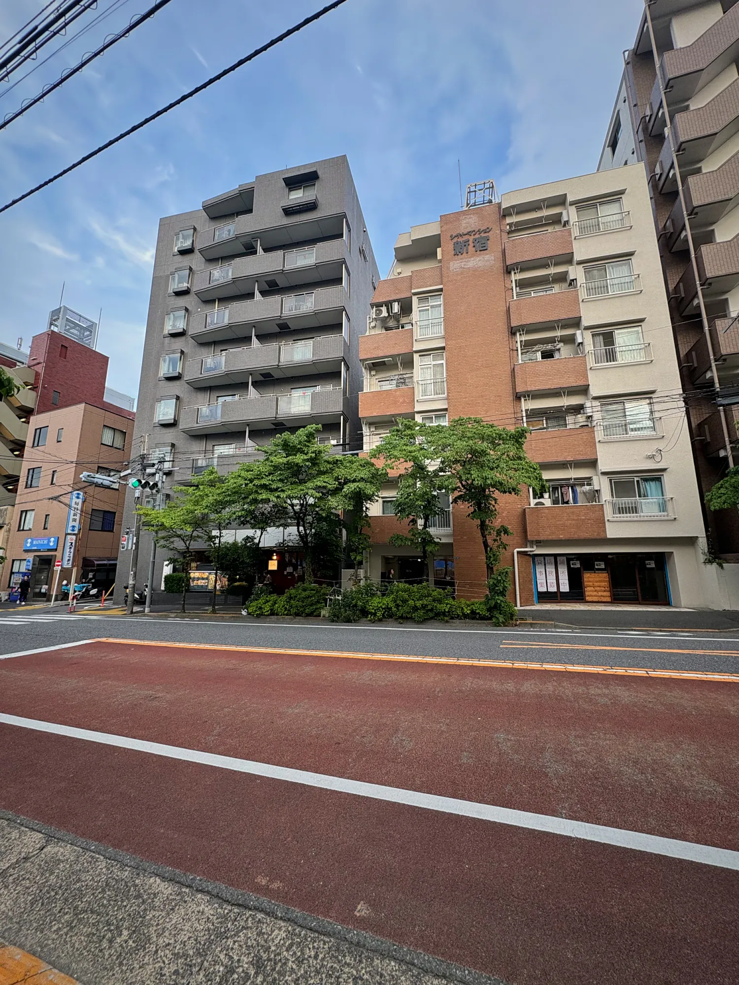 A quiet residential street in Shinjuku with apartment buildings, green trees, and a red-paved bike lane.