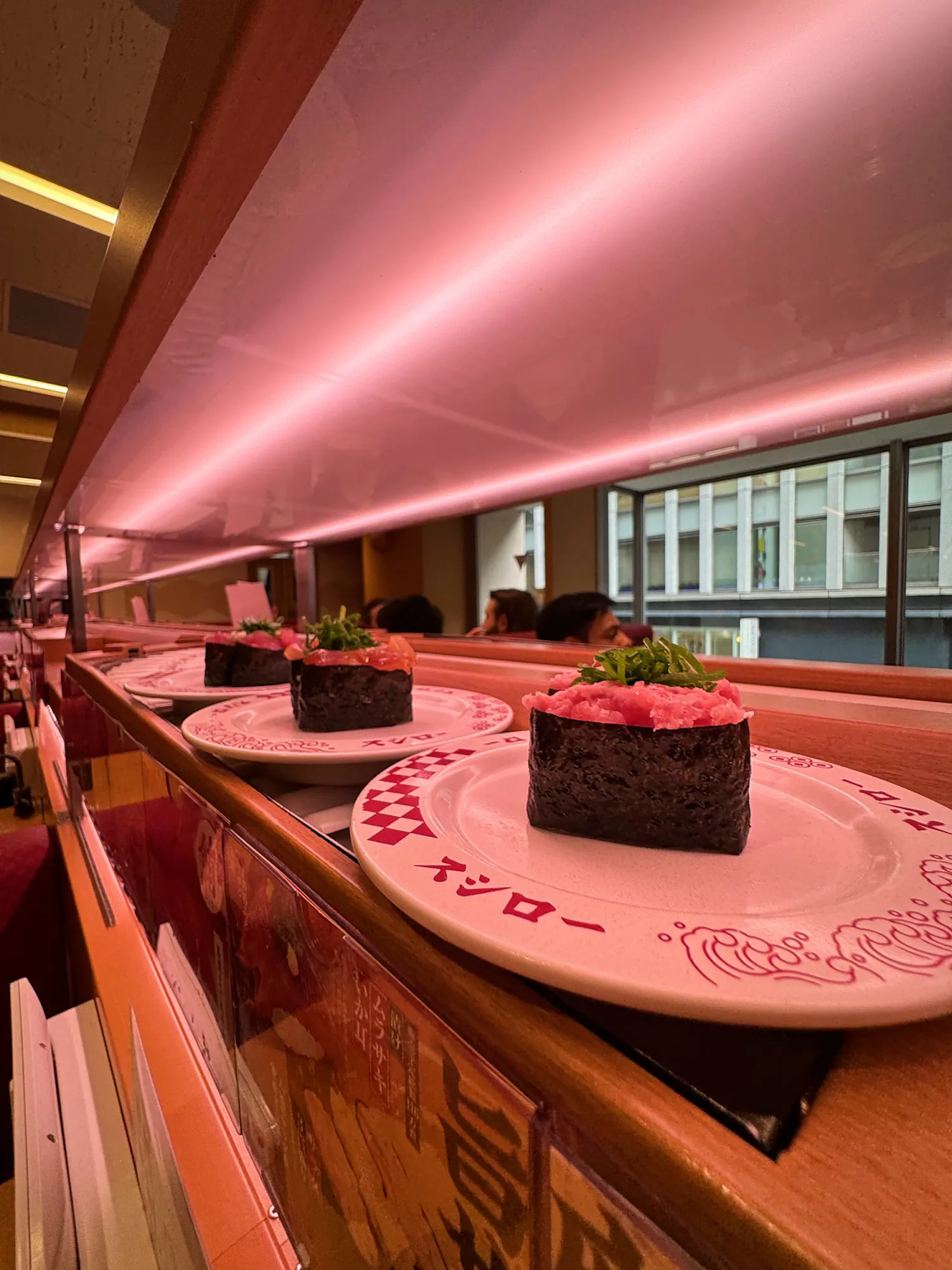 Gunkan sushi topped with tuna and green onion on Sushiro plates, riding the pink-lit express lane in Tokyo.