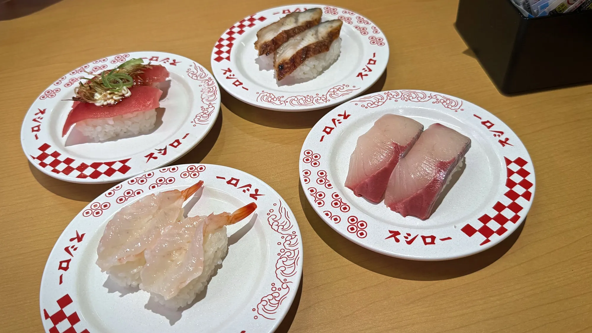 Four plates of nigiri sushi on a wooden table at Sushiro in Tokyo, including tuna, yellowtail, shrimp, and grilled fish.