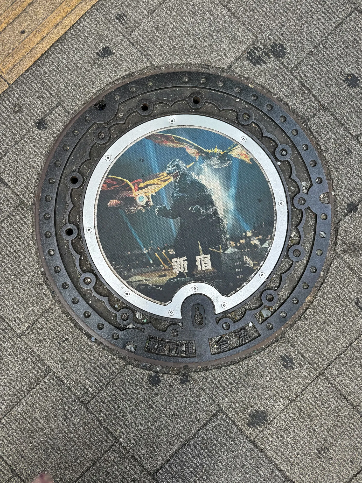 A round manhole cover with a full-color Godzilla design set into a Shinjuku sidewalk.