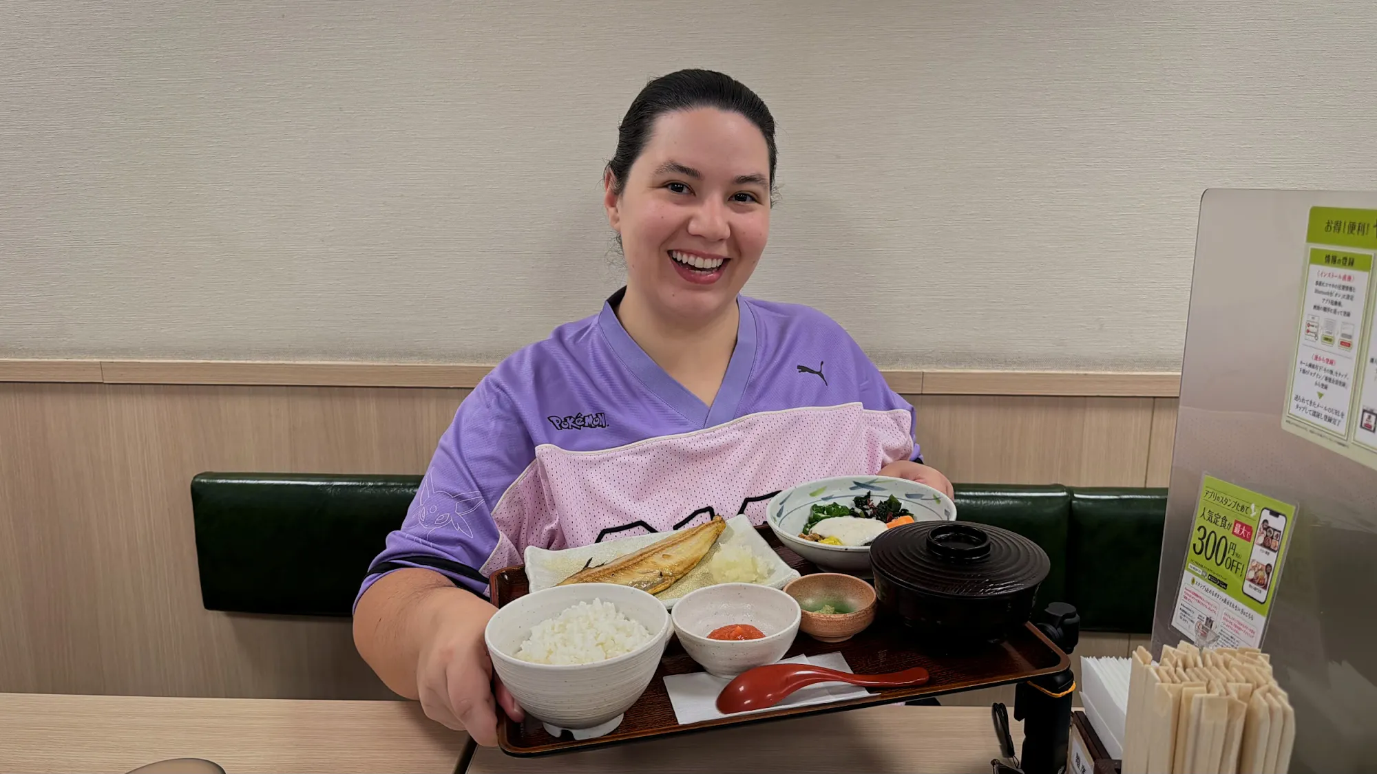 A smiling woman in a purple top holding a Japanese breakfast tray at Yayoiken in Tokyo.