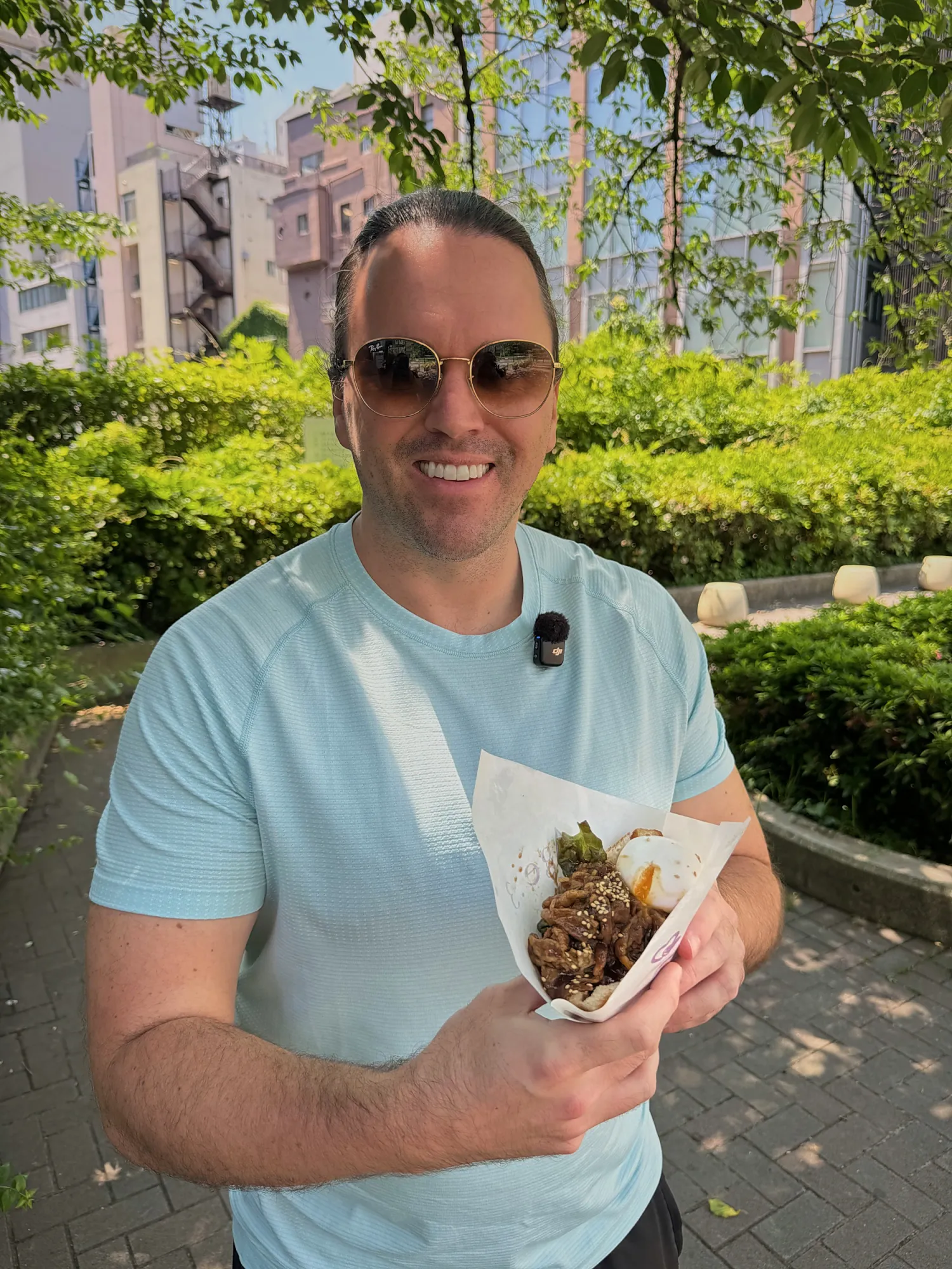 Cream smiles in a light blue T-shirt and sunglasses while holding street food beside a leafy hedge in Tokyo.