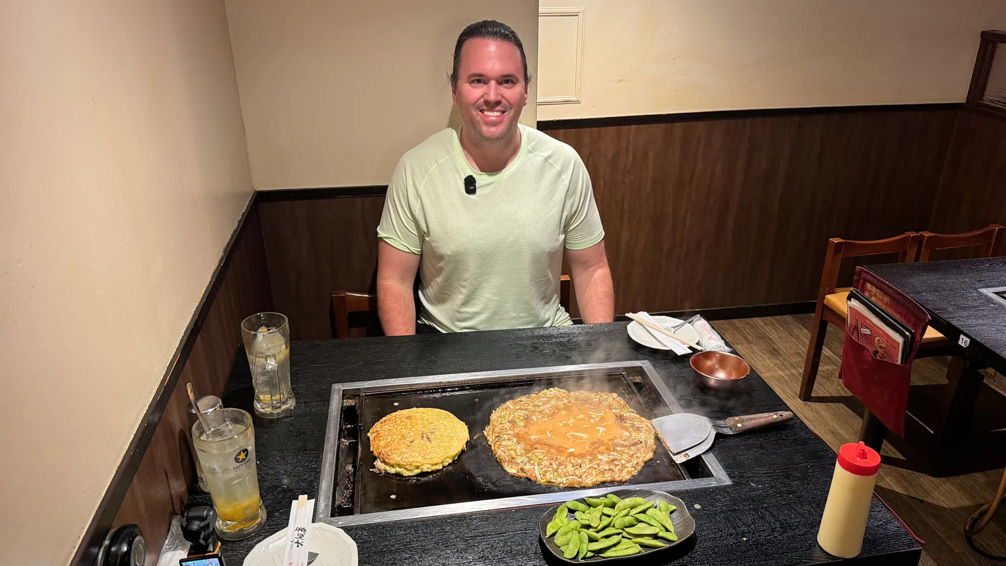 Cream grinning behind a griddle with okonomiyaki and monjayaki cooking and edamame on the side at Osakaya