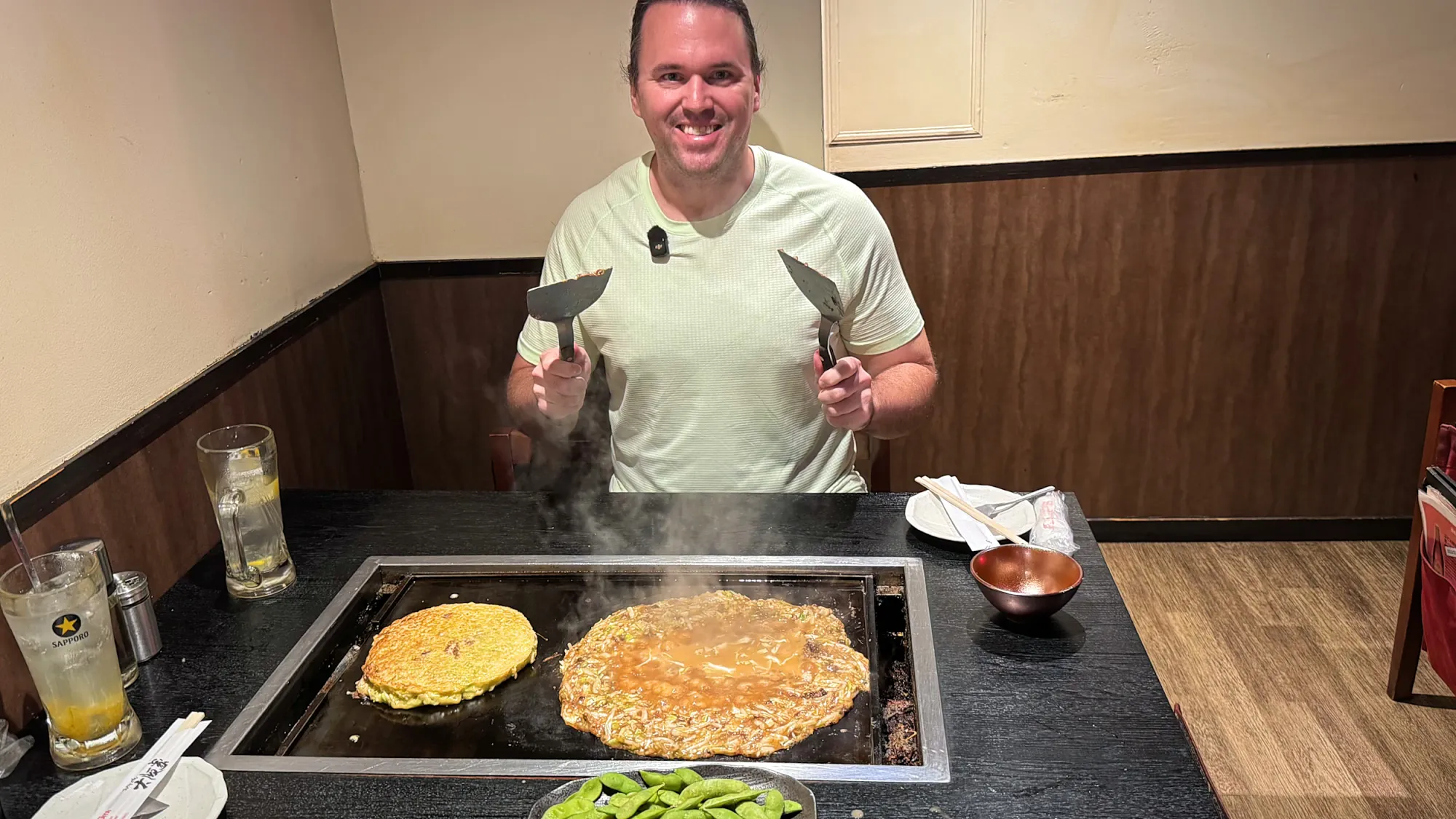 Cream smiling and holding two metal spatulas over a table griddle with okonomiyaki and monjayaki cooking at Osakaya in Shinjuku, Tokyo.
