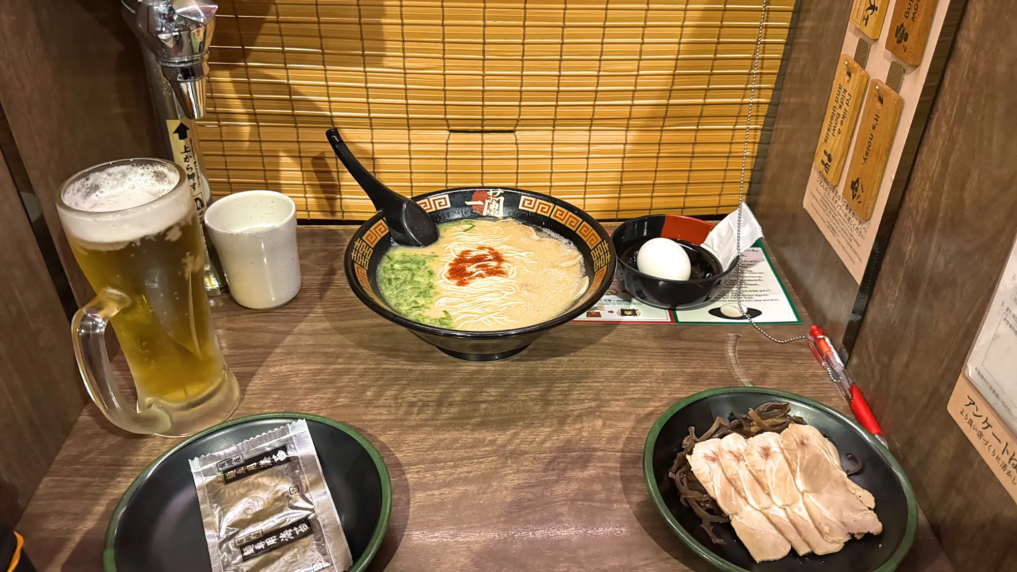 An Ichiran tonkotsu ramen set in a wooden solo booth with a draft beer, a boiled egg, and a plate of sliced pork