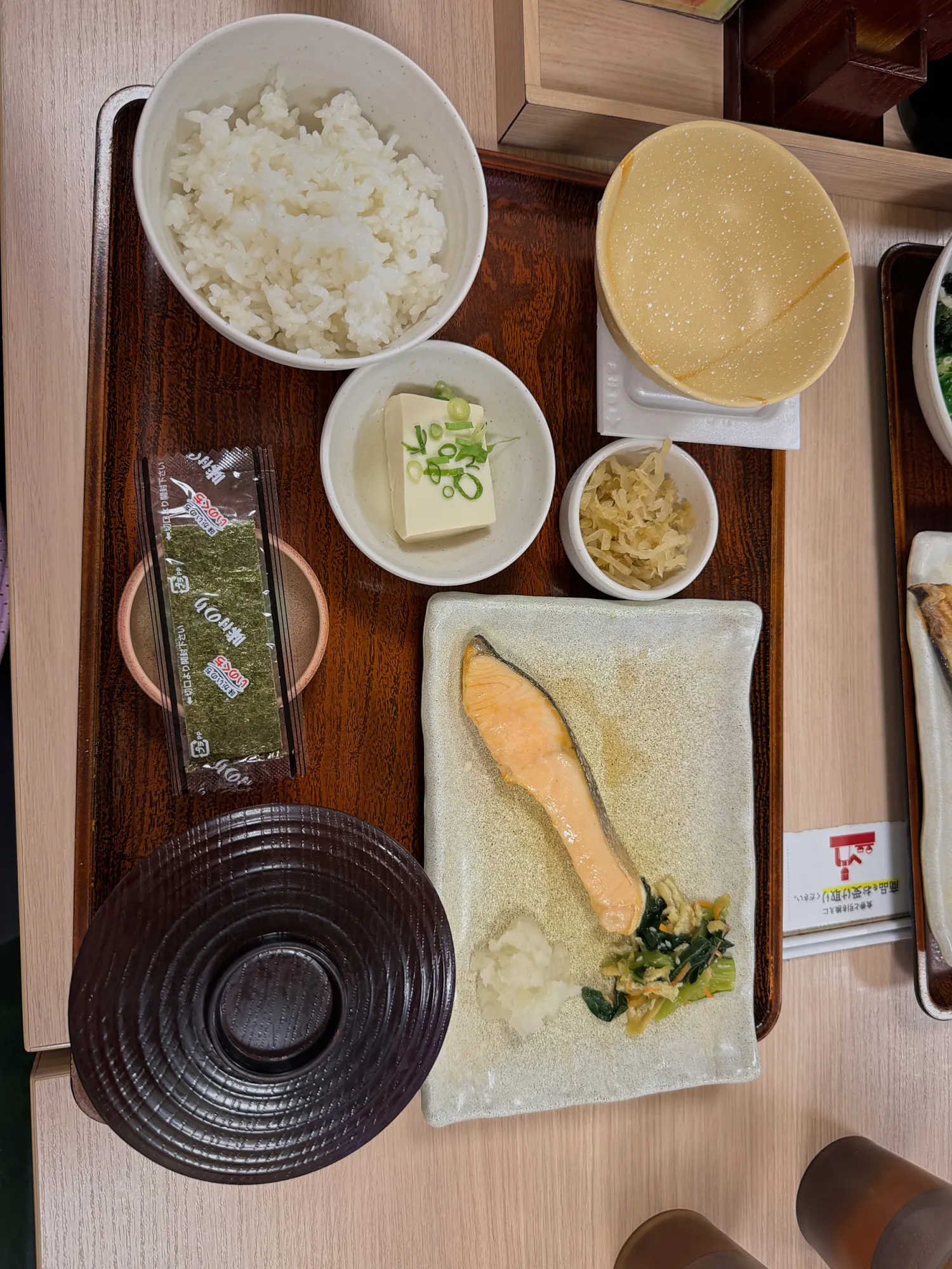 A Japanese breakfast set at Yayoiken in Tokyo seen from above: grilled salmon with greens and grated daikon, rice, chilled tofu with scallions, natto, pickles, a packet of nori, and miso soup with its lid on.