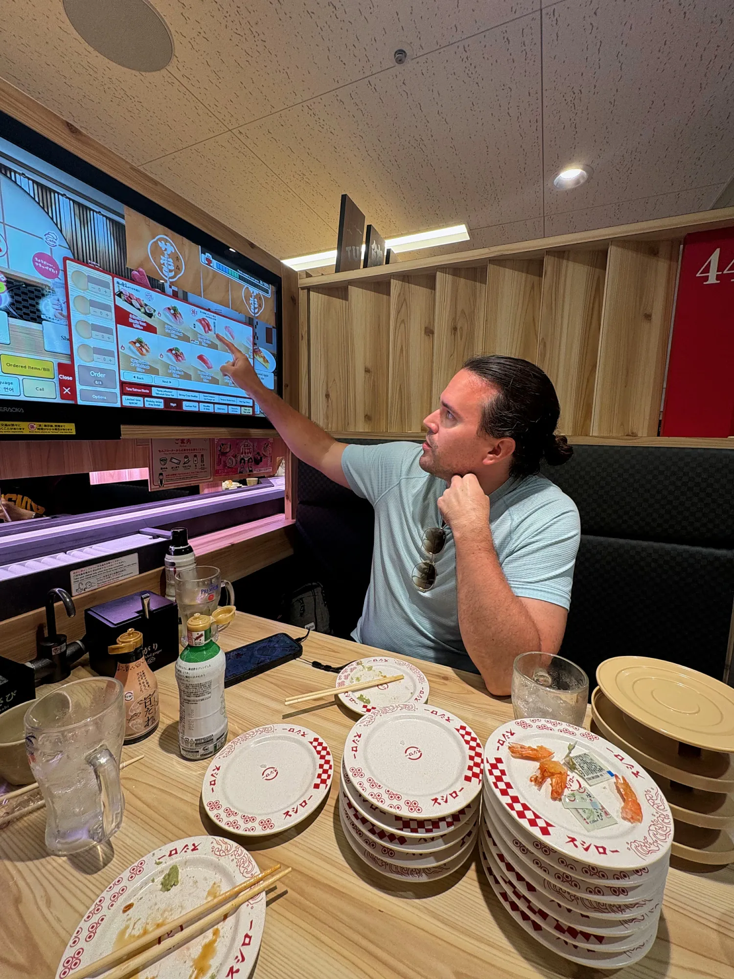 Cream pointing at a large wall-mounted ordering screen above a table stacked with empty Sushiro plates in Tokyo.