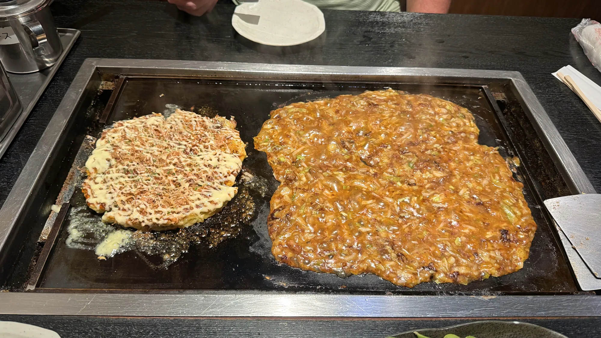 An okonomiyaki and a spread of monjayaki cooking side by side on the table griddle at Osakaya