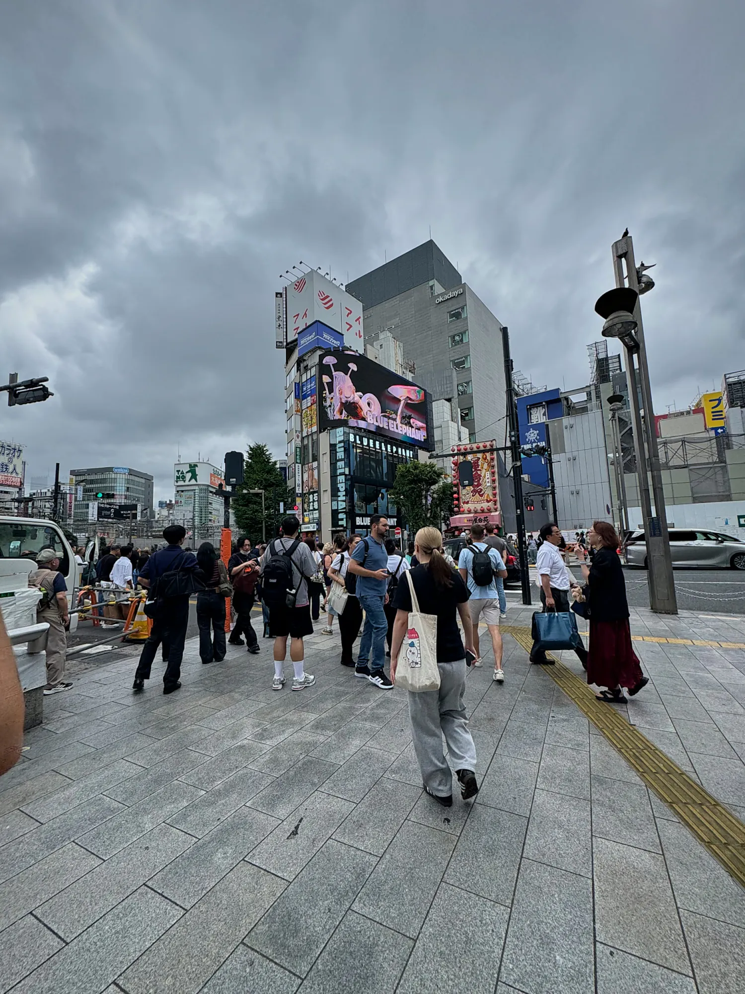 A wide pedestrian plaza outside Shinjuku Station in Tokyo, with people walking under a cloudy sky and billboards on the buildings ahead.