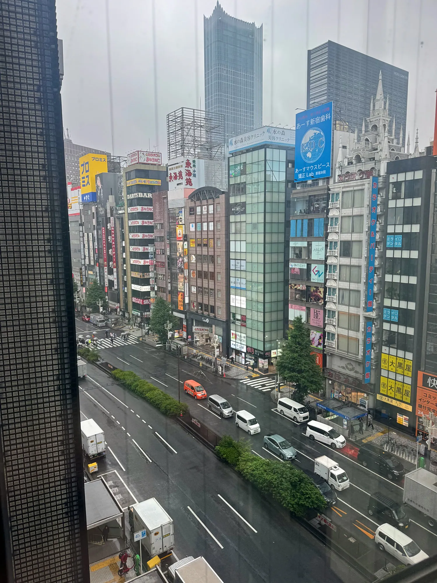 A wet, gray Shinjuku avenue seen from a window above, with cars on the road and tall buildings under heavy cloud.