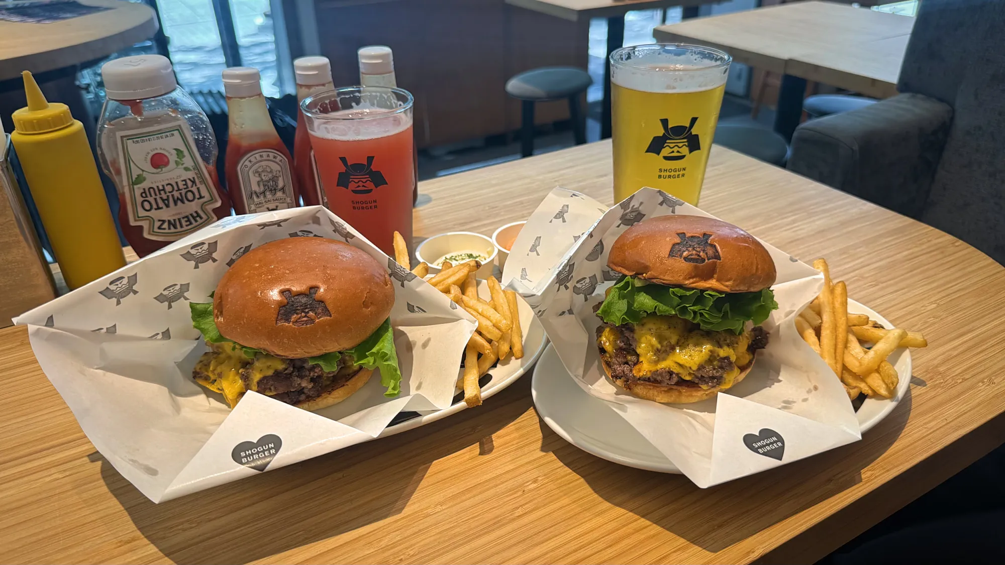 Two Shogun Burger wagyu cheeseburgers with fries side by side on a table, with sauces and a beer behind