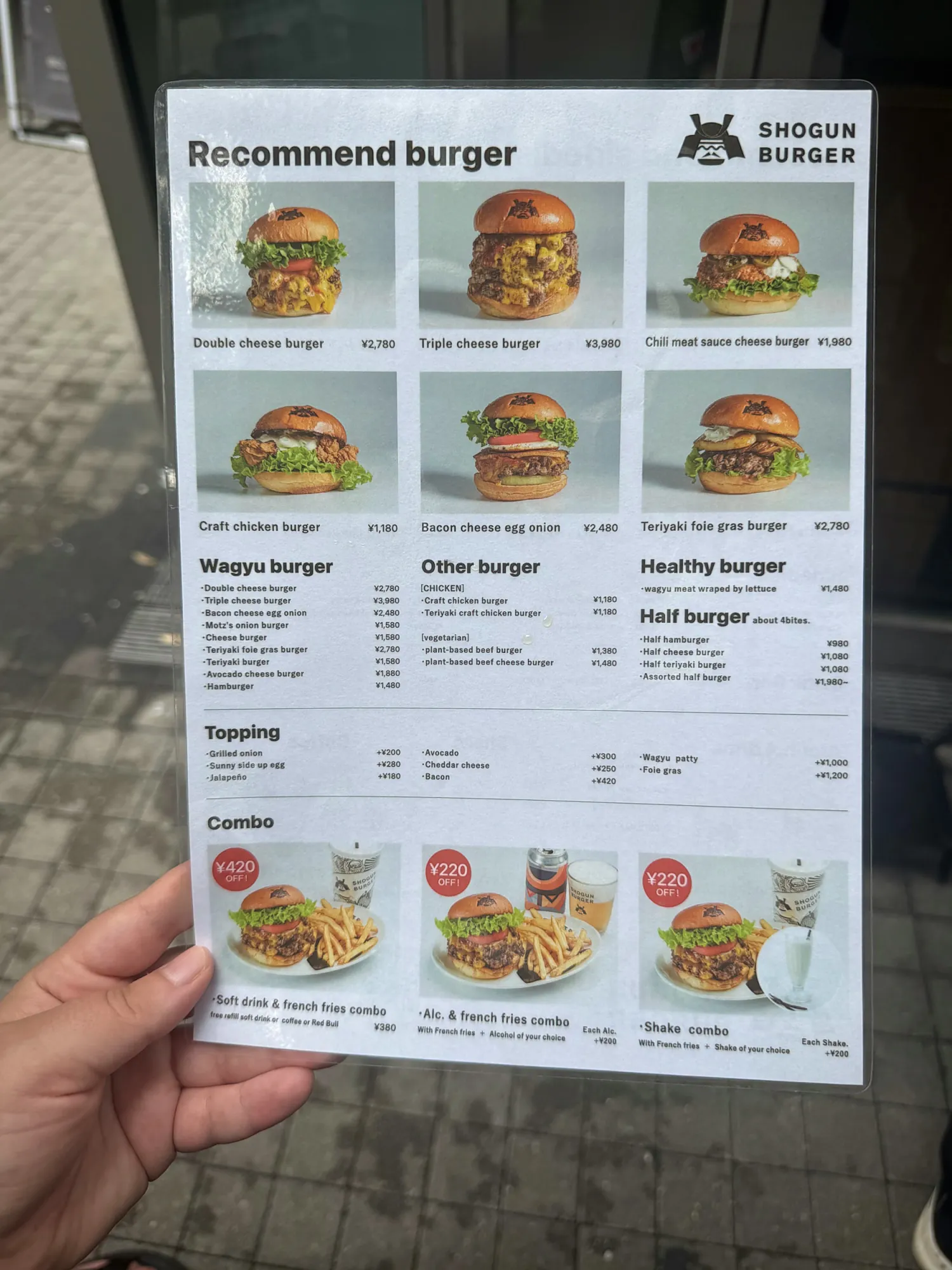 A hand holding the English Shogun Burger menu in Tokyo, with photos and prices of the double cheese burger, triple cheese burger, chicken burger, and combo deals.