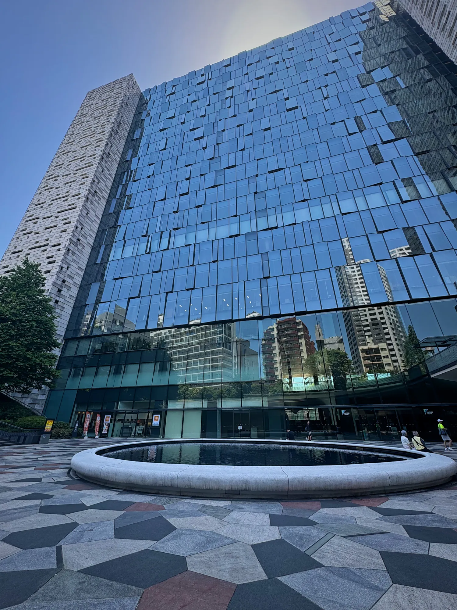 A glass office tower reflecting the Tokyo sky above a round fountain on a patterned stone plaza.