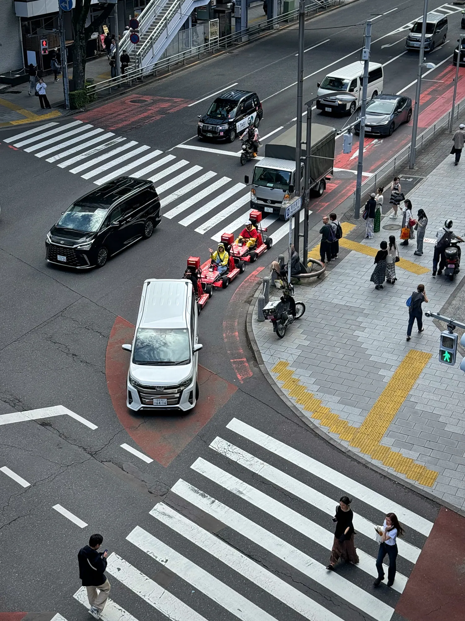 A line of street go-karts with costumed drivers turning through an intersection between cars in Tokyo, seen from above.