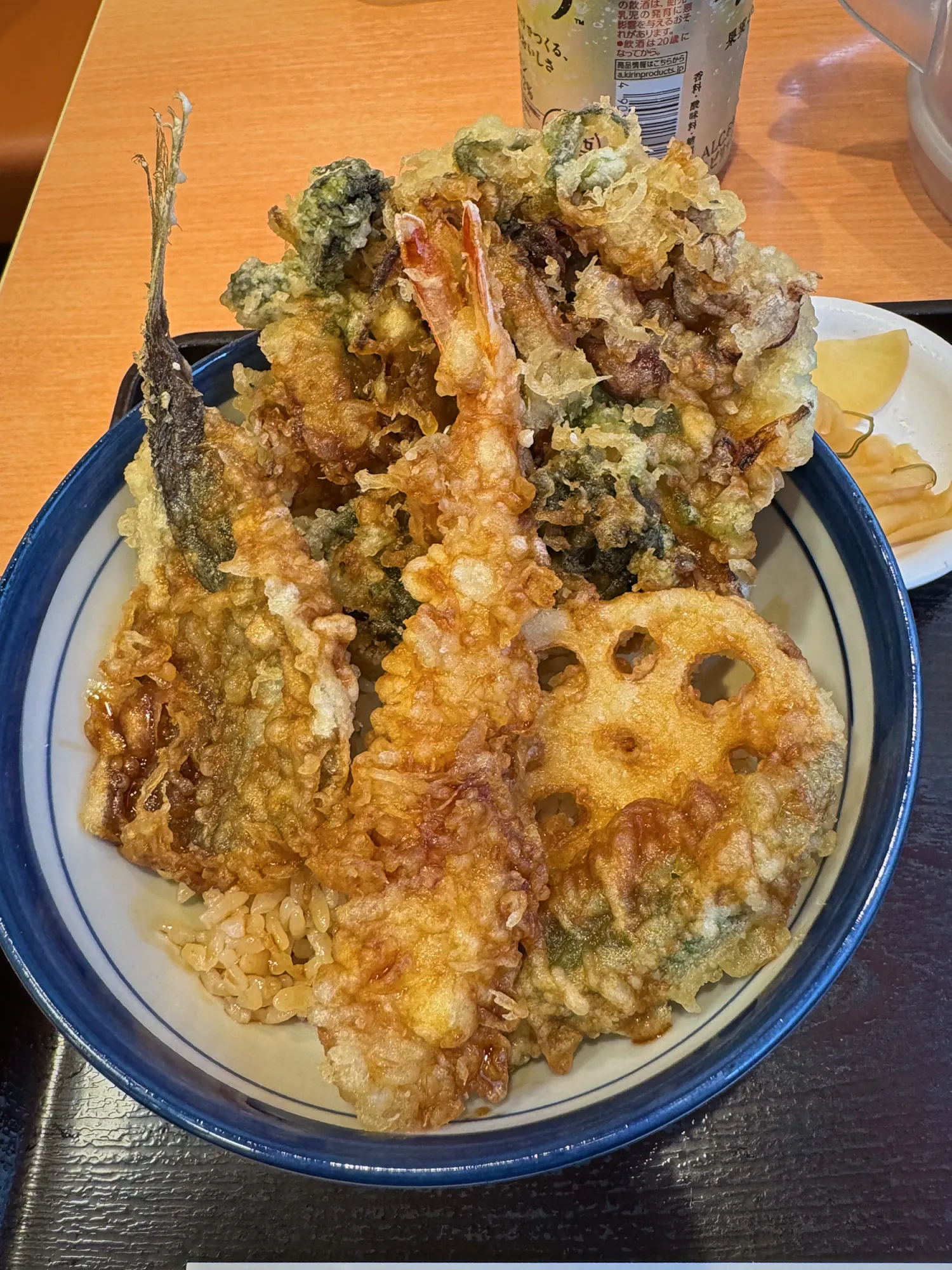 A Tendon Tenya tempura bowl piled with shrimp, a whole small fish, lotus root, and a vegetable fritter over sauce-soaked rice, with yellow pickles on the side.
