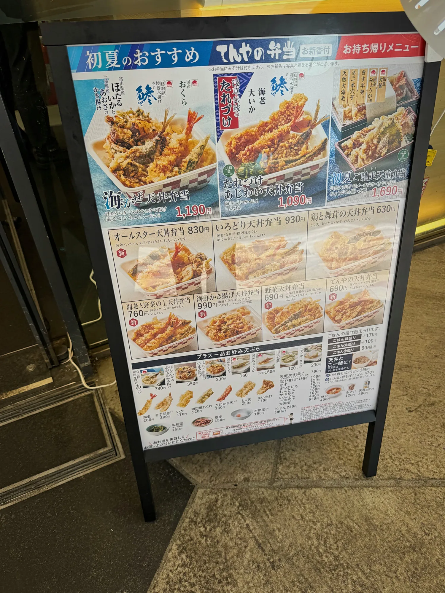 A Tendon Tenya takeaway menu board in Japanese outside the shop, showing tempura bento boxes and their prices in yen.