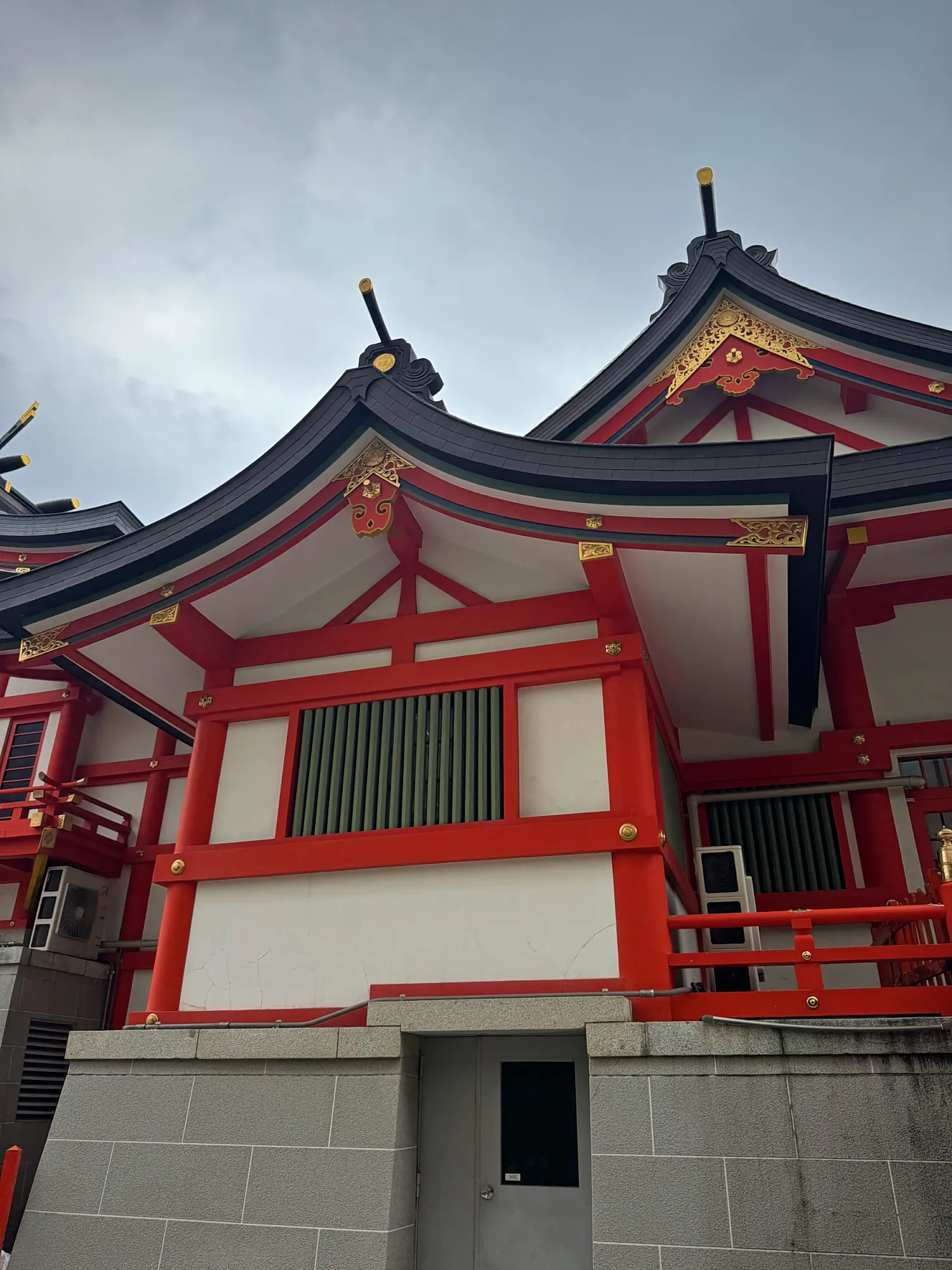 Red and white wooden shrine buildings with curved black roofs under a cloudy sky in Shinjuku, Tokyo.
