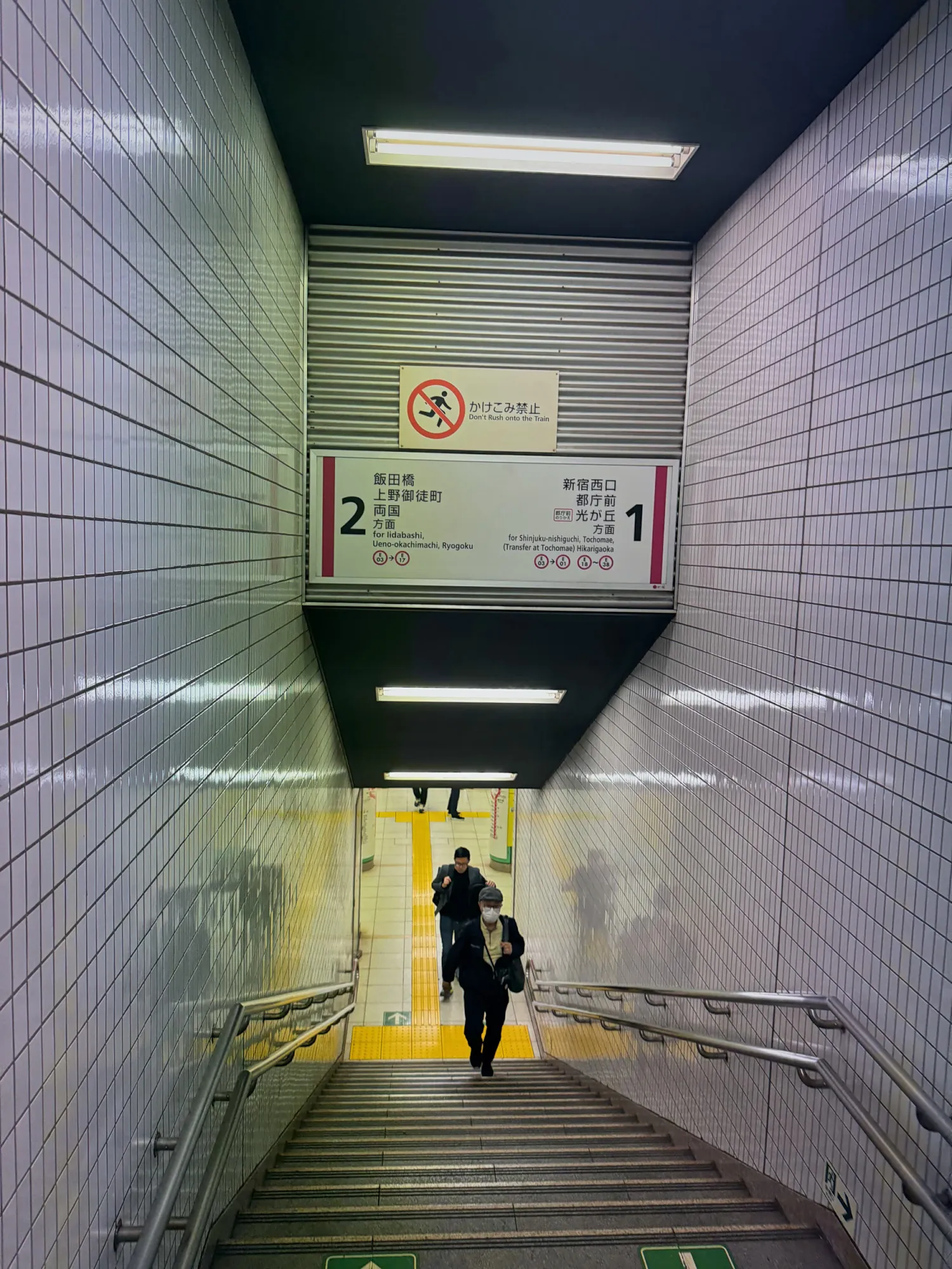 A tiled Tokyo subway staircase with signs numbering each side, as a traveler walks down to the platform.