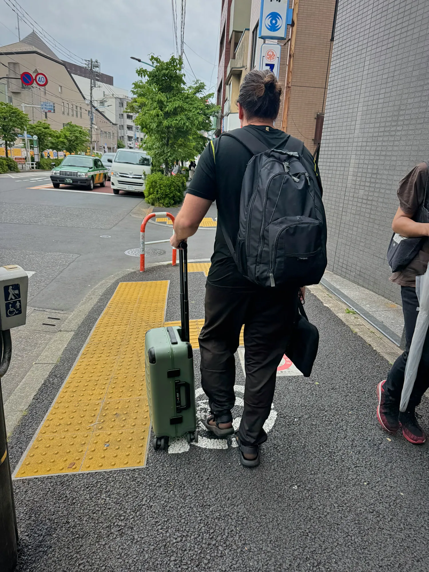 A traveler with a backpack pulling a green suitcase along a quiet sidewalk in a residential part of Shinjuku, Tokyo.