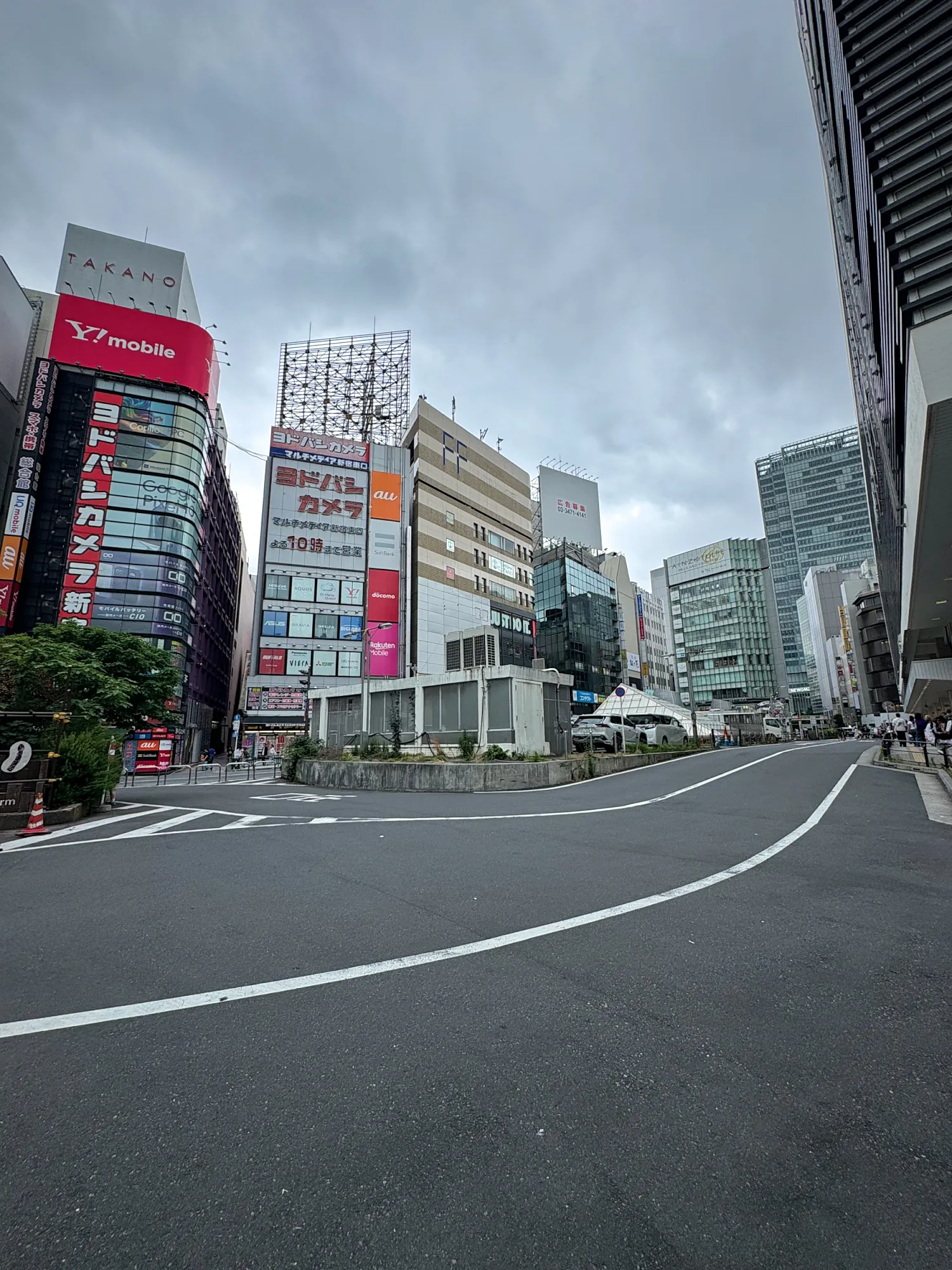 A wide, quiet Shinjuku street and crosswalk under a flat gray sky, with tall buildings and shop signs on both sides.