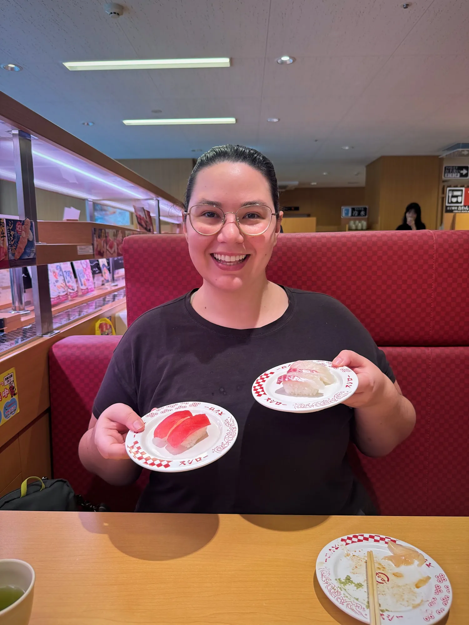 Bernie smiling in a red Sushiro booth in Tokyo, holding up two plates of nigiri.