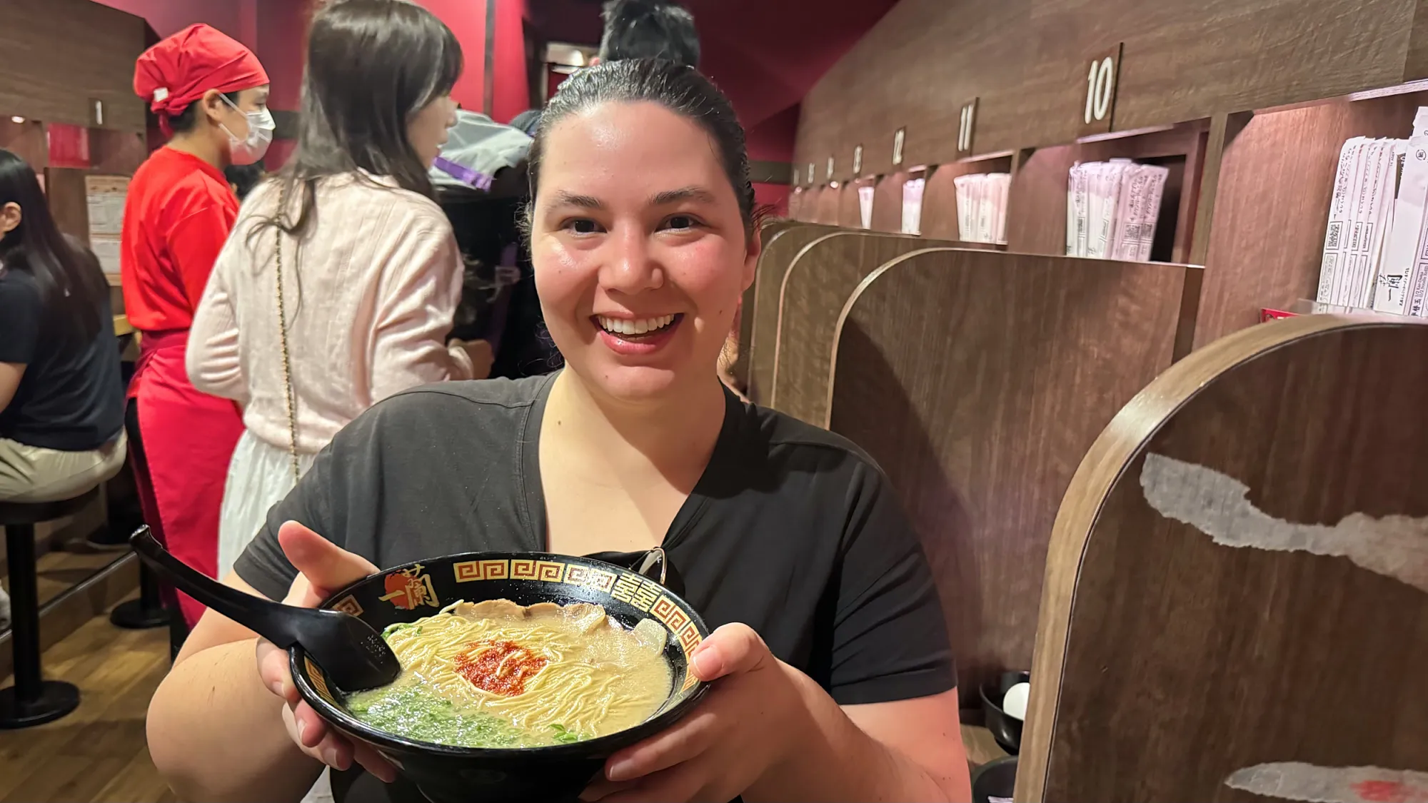 Bernie smiling with her bowl of Ichiran ramen in a solo booth, with diners and staff in red filling the room behind her.