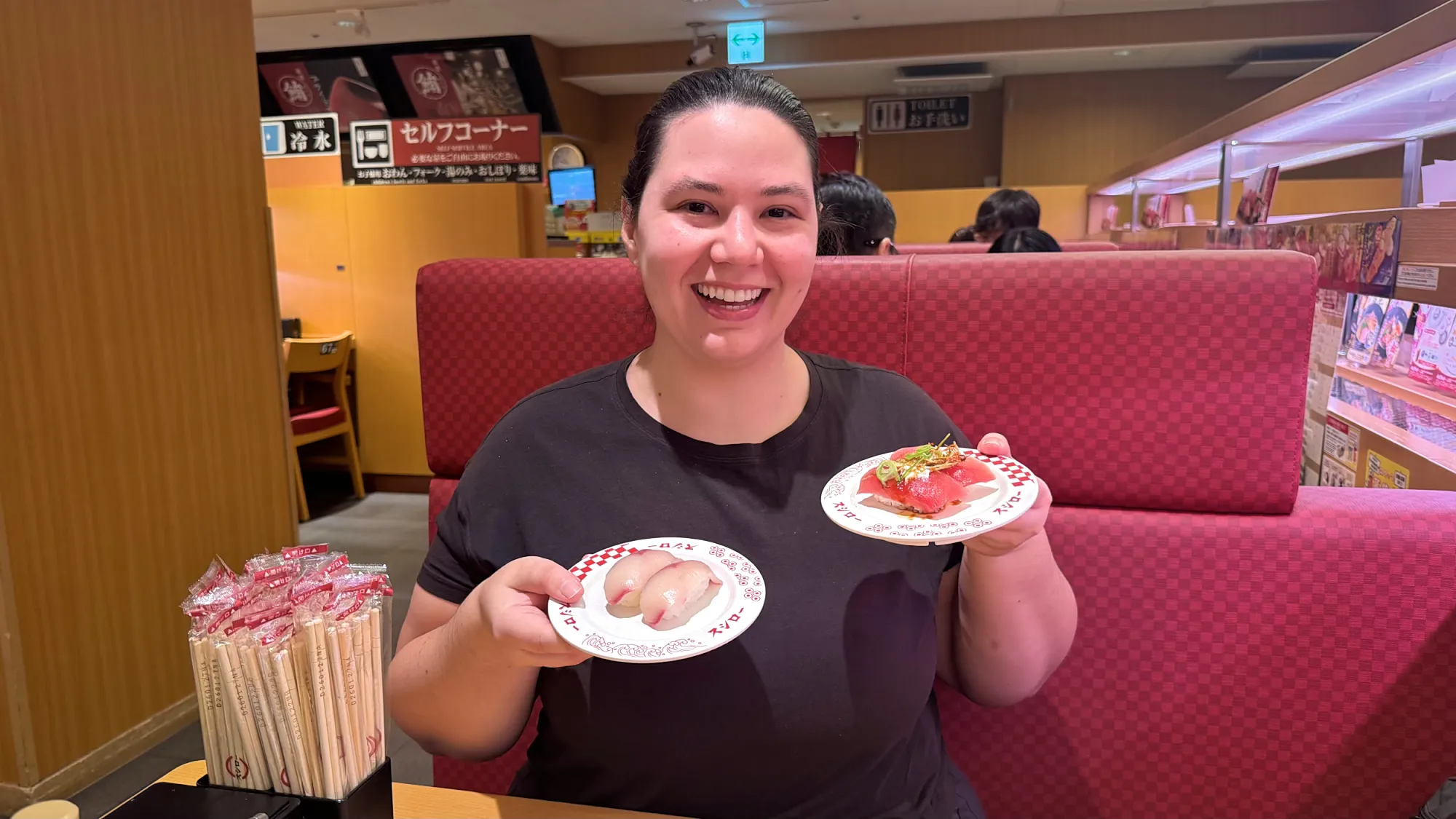 Bernie smiling in a red Sushiro booth, holding up two plates of sushi