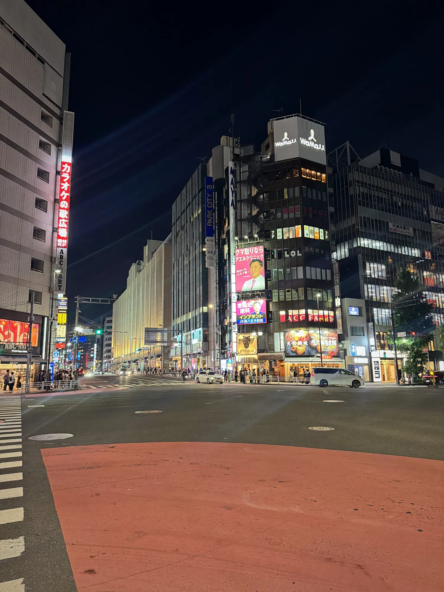 A wide Shinjuku intersection at night, with lit-up buildings and signs around an empty crosswalk.