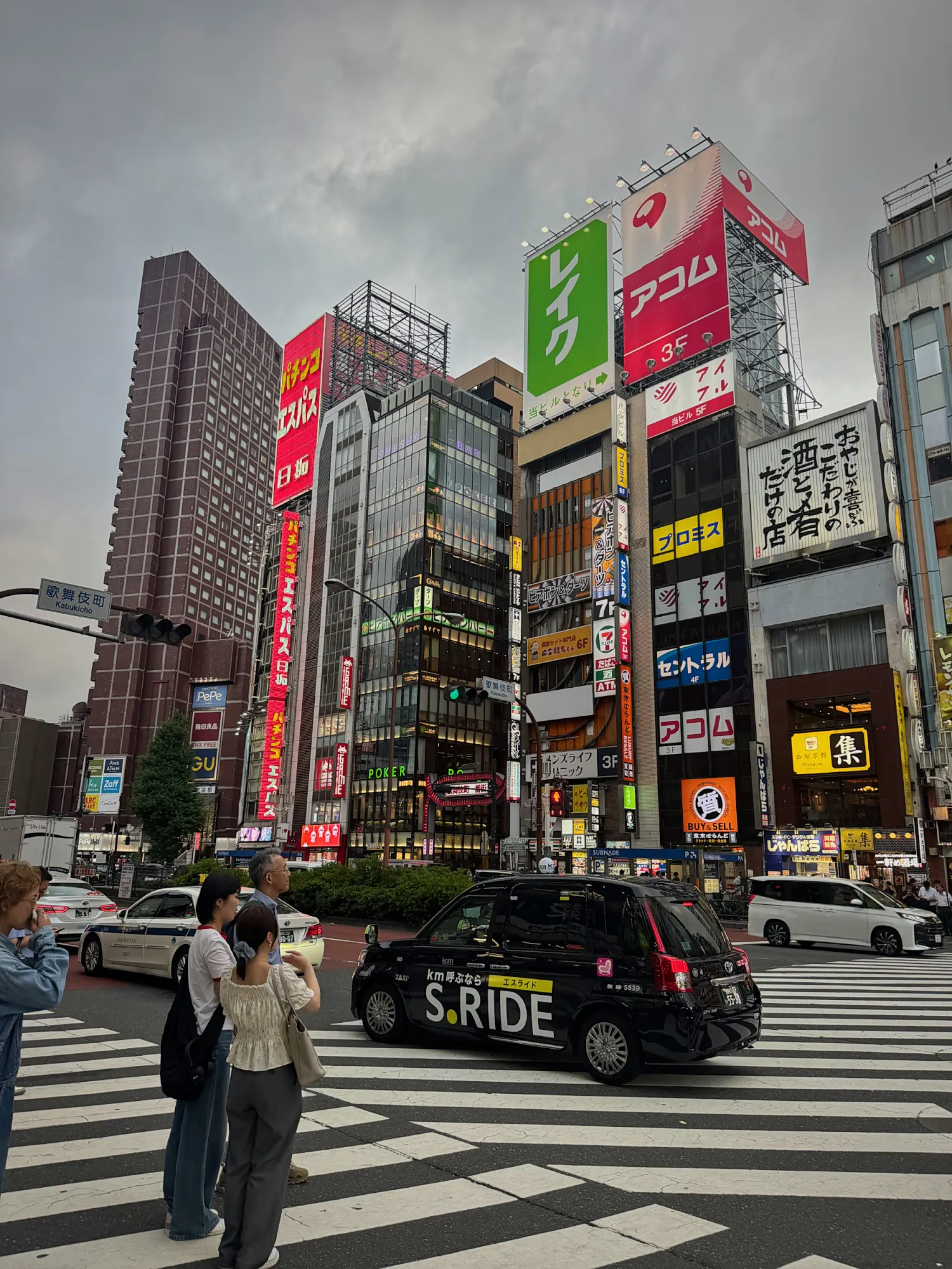 A Shinjuku intersection at night under tall buildings covered in neon signs, with a taxi waiting at the crosswalk, Tokyo.