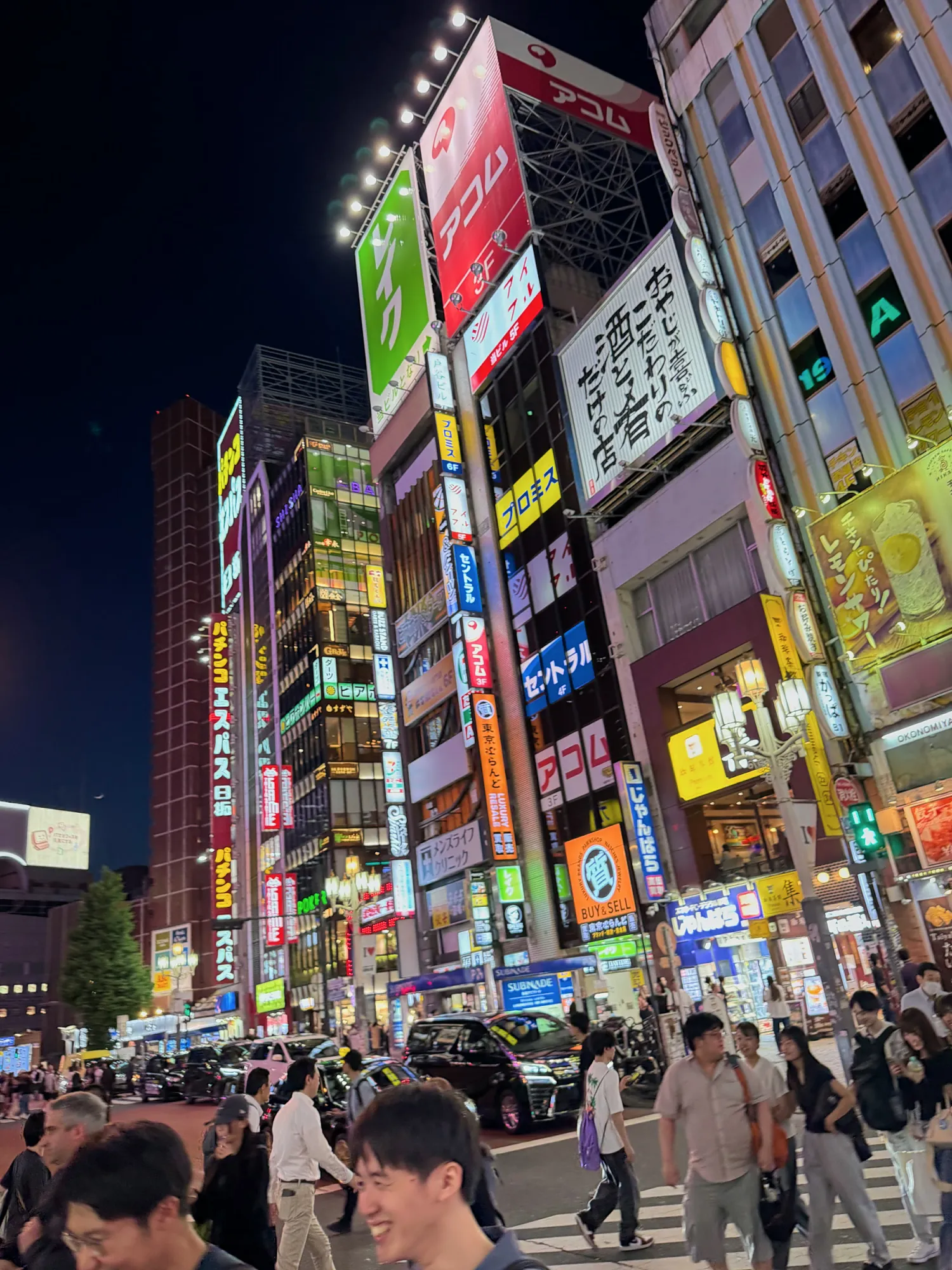 People crossing a Shinjuku street at night under towering neon signs and bright storefronts.
