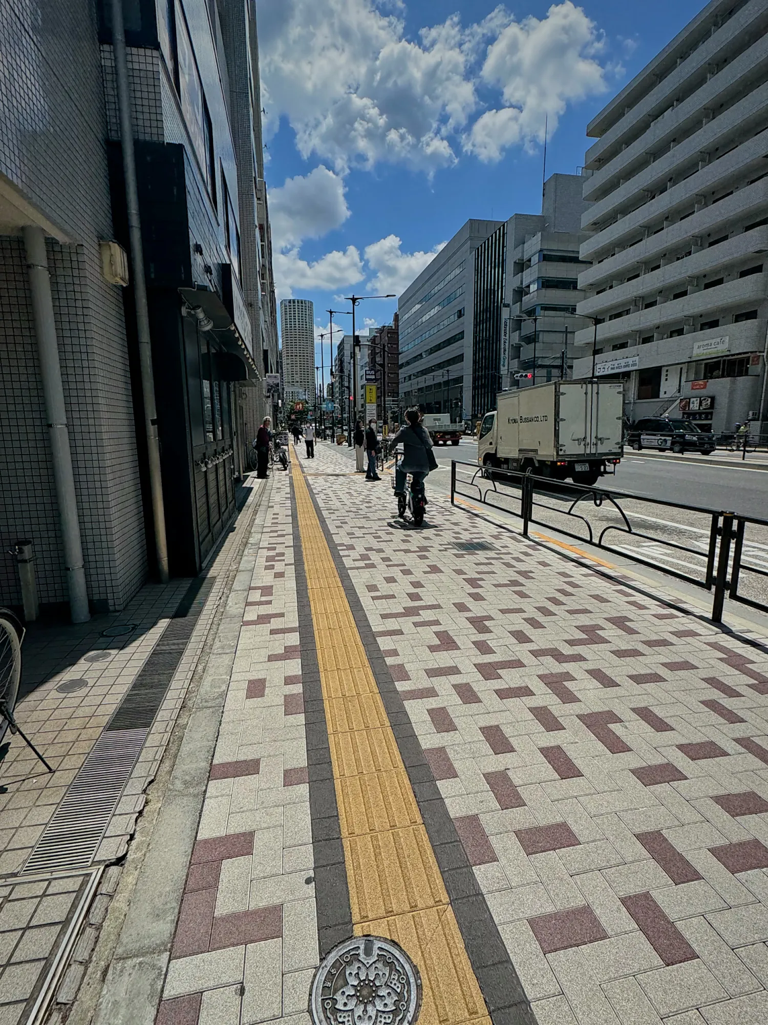 A clean, wide sidewalk with yellow tactile paving and no benches along a street in Shinjuku, Tokyo.