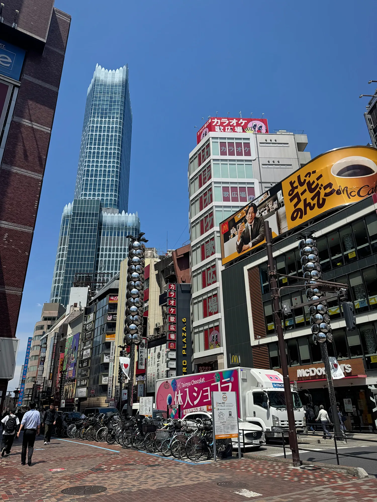 Shops and signs along a busy Shinjuku street in Tokyo, with the tall Tokyu Kabukicho Tower rising behind them against a clear blue sky.
