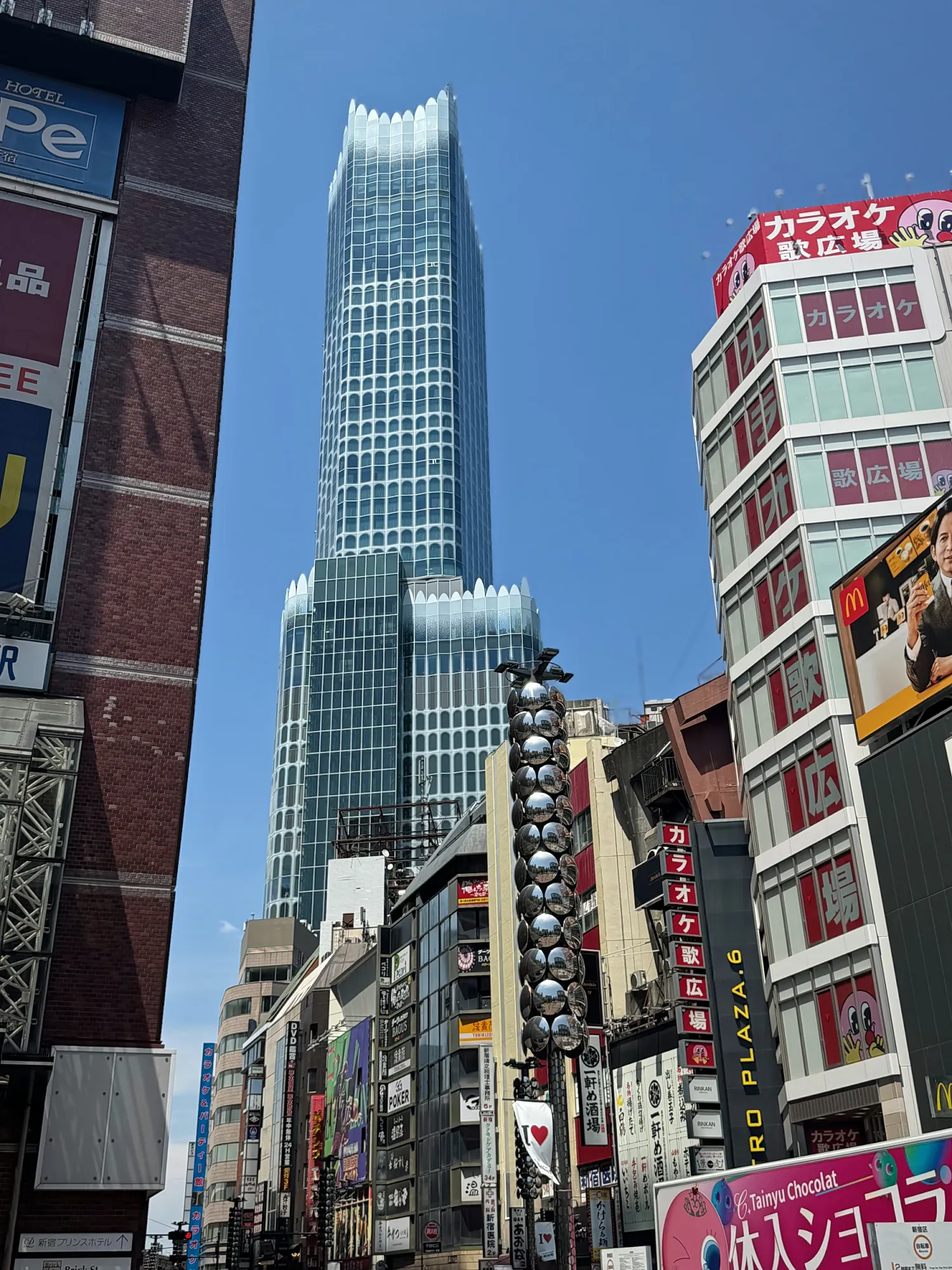 Tokyu Kabukicho Tower rising above karaoke signs and shop buildings on a street in Shinjuku, Tokyo, under a clear blue sky.