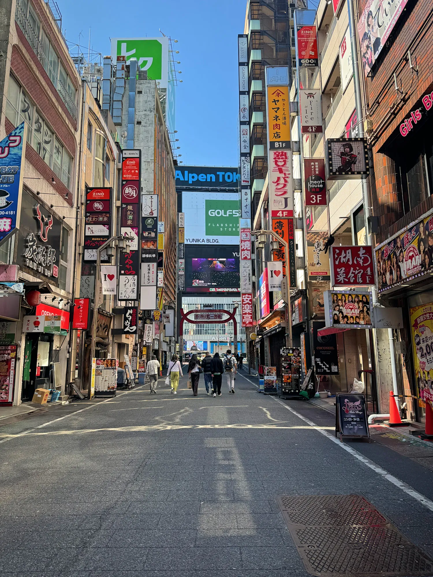 A Kabukicho street in Shinjuku, Tokyo, lined with tall vertical shop signs under a clear blue morning sky.