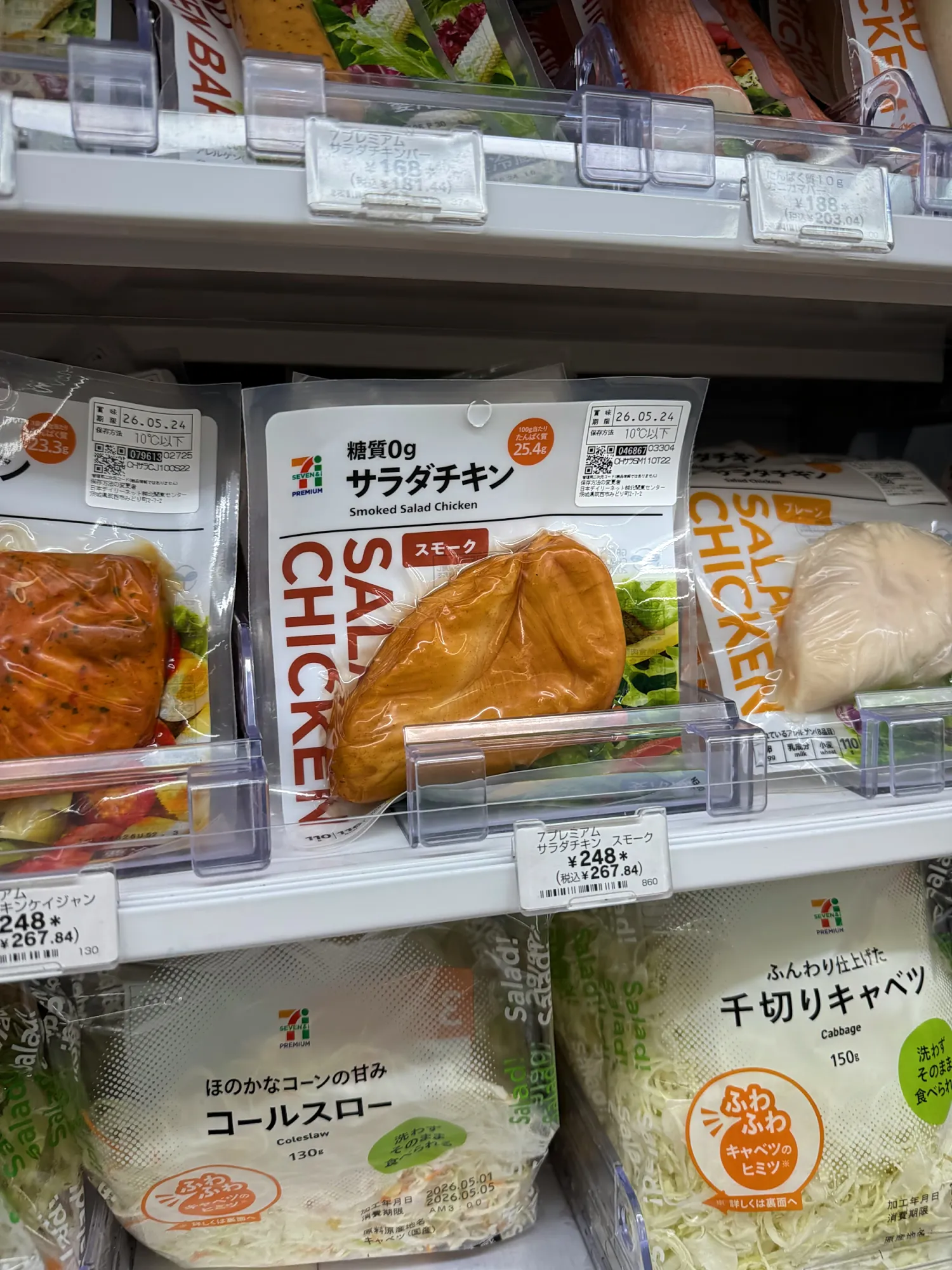 A 7-Eleven fridge shelf in Tokyo with packs of pre-cooked smoked chicken breast labeled 0 g sugar at 248 yen, above bags of shredded cabbage and coleslaw.