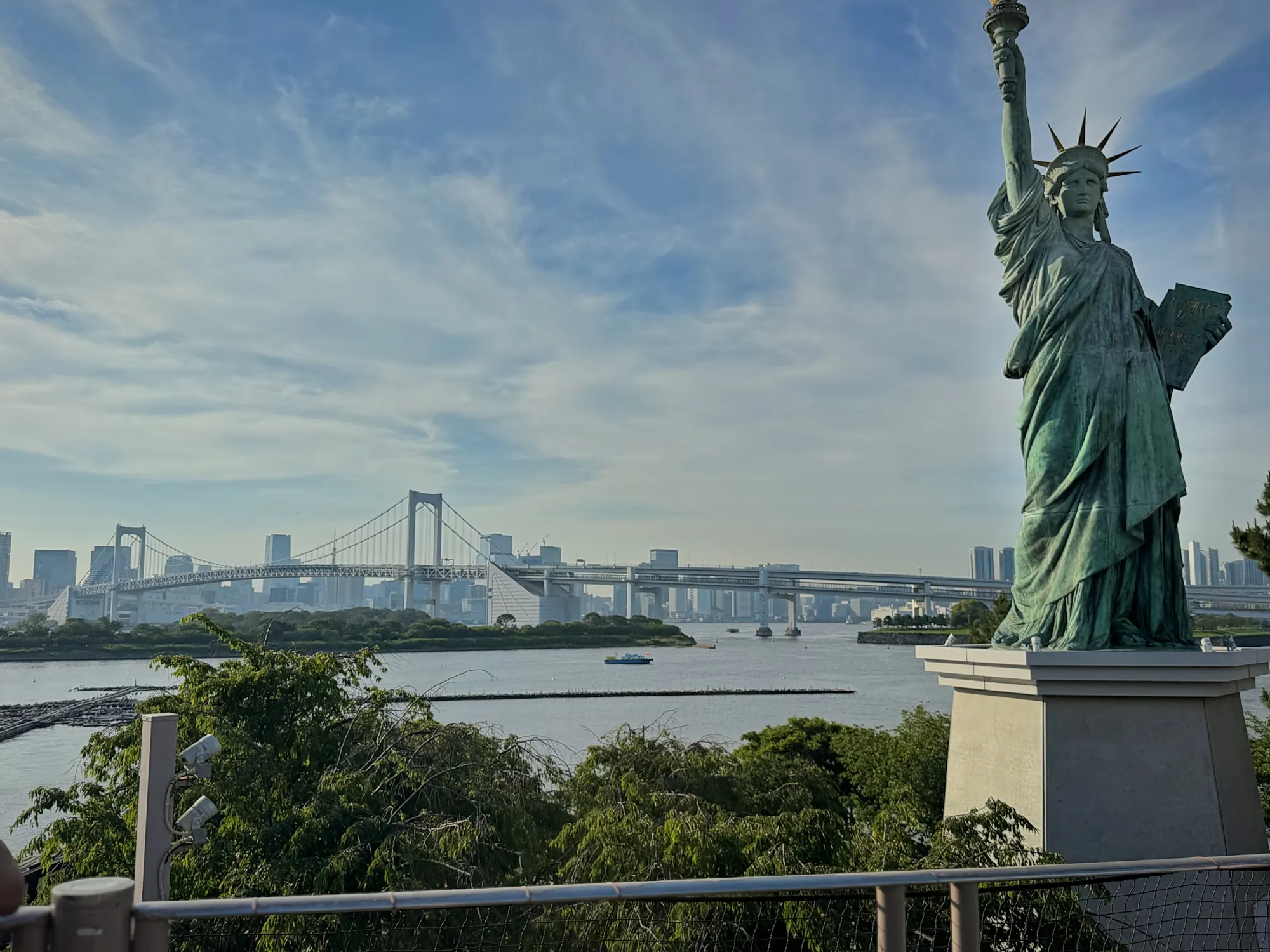 The Statue of Liberty replica in Odaiba, Tokyo, with Rainbow Bridge and Tokyo Bay behind it under a blue sky.