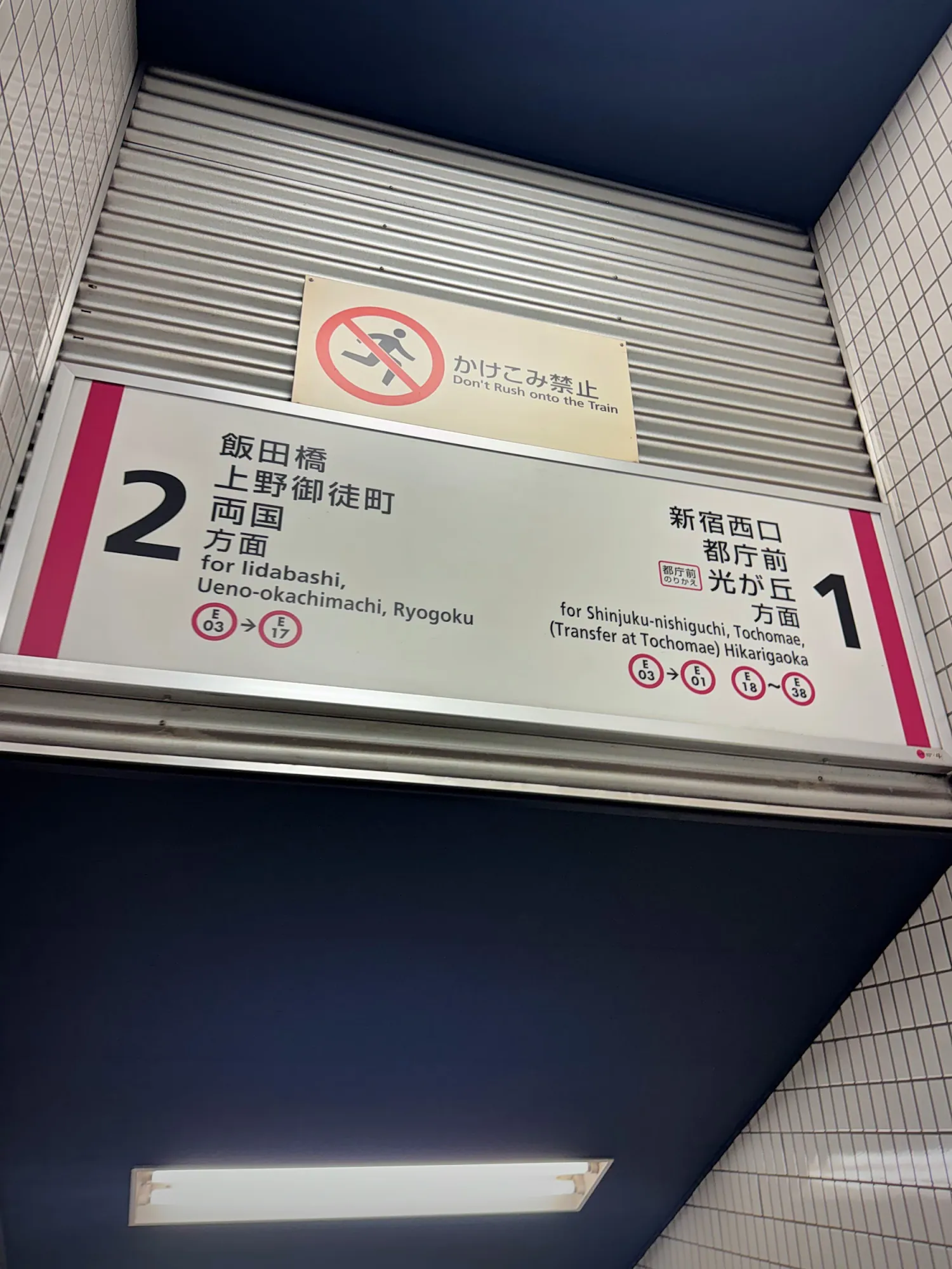 A sign above a stairway in a Tokyo subway station showing platform 2 to the left for Iidabashi, Ueno-okachimachi, and Ryogoku, and platform 1 to the right for Shinjuku-nishiguchi.