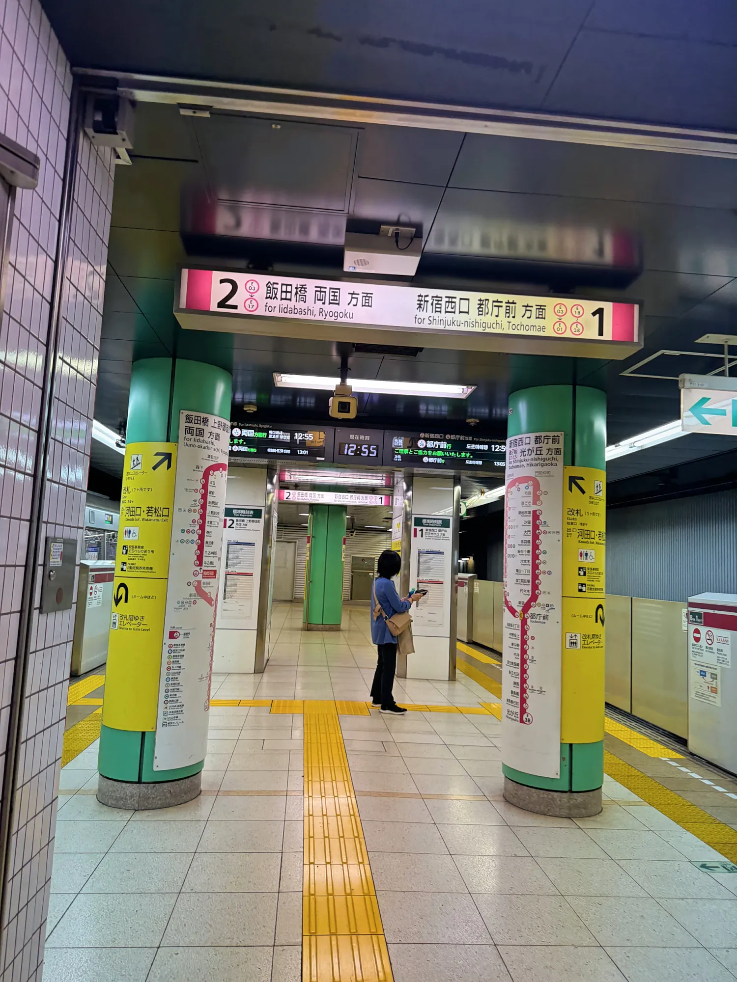 A clean Toei Oedo Line platform at Wakamatsu-kawada Station in Tokyo, with signs for platform 2 toward Iidabashi and platform 1 toward Shinjuku-nishiguchi and route maps on the pillars.